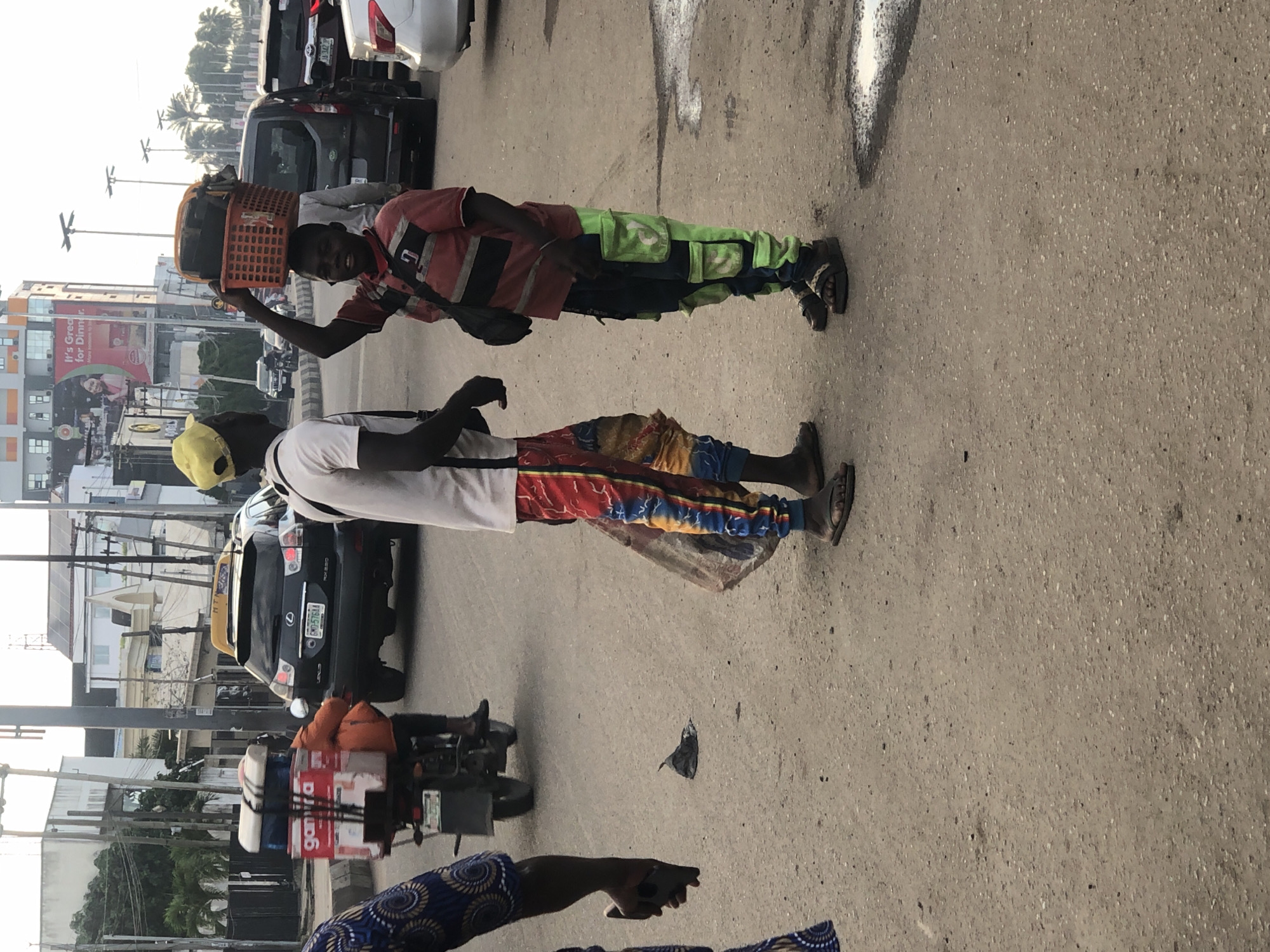In Ikeja, Nigeria, on a paved road, a young Black male walks with an orange basket filled with items on his head. He wears a red and black striped shirt and dark pants with bright green pockets. Beside him, another Black male, wearing a yellow cap, white t-shirt, and colorful patterned pants, stands and faces him. A motorcycle with items strapped to the back is visible to the left, and several cars are present in the background, including a black Lexus SUV and a white car. Buildings with signage, including a billboard advertising "It's Great for Dinner," are in the distance, along with streetlights and palm trees. The ground is dusty and there are some water puddles.