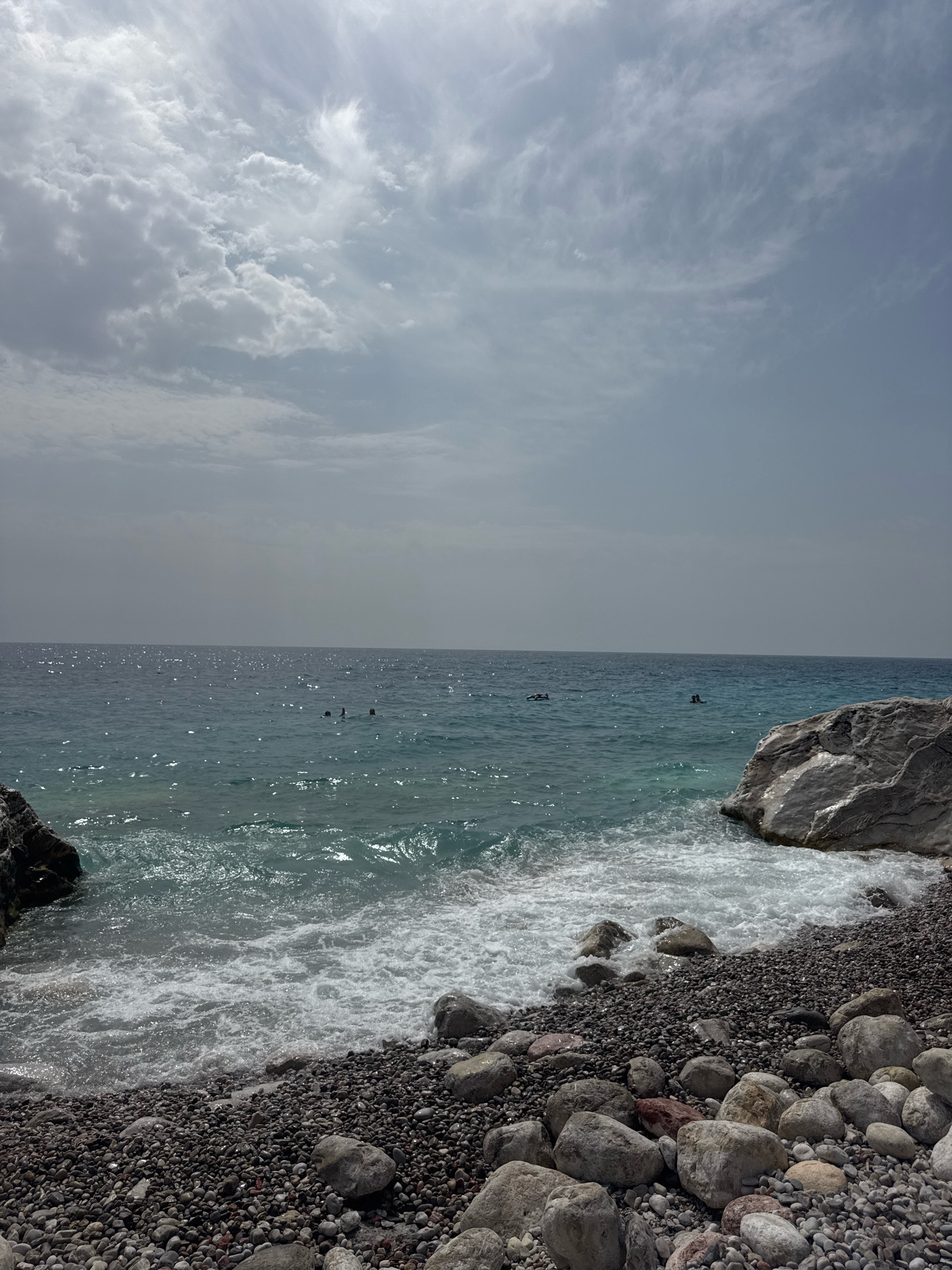 The image depicts a rocky beach on a sunny day with a partially cloudy sky. The turquoise water gently laps onto the shore, creating white foam. In the distance, several people are visible swimming or floating in the sea. The beach is covered in smooth, dark pebbles and larger grey rocks. The sky is a pale blue with scattered white clouds, some of which are wispy. The scene appears to be a natural coastal area, likely a beach or cove. The provided crops highlight the texture of the clouds, the shimmer of sunlight on the water, and the detail of the rocks and pebbles on the shore.