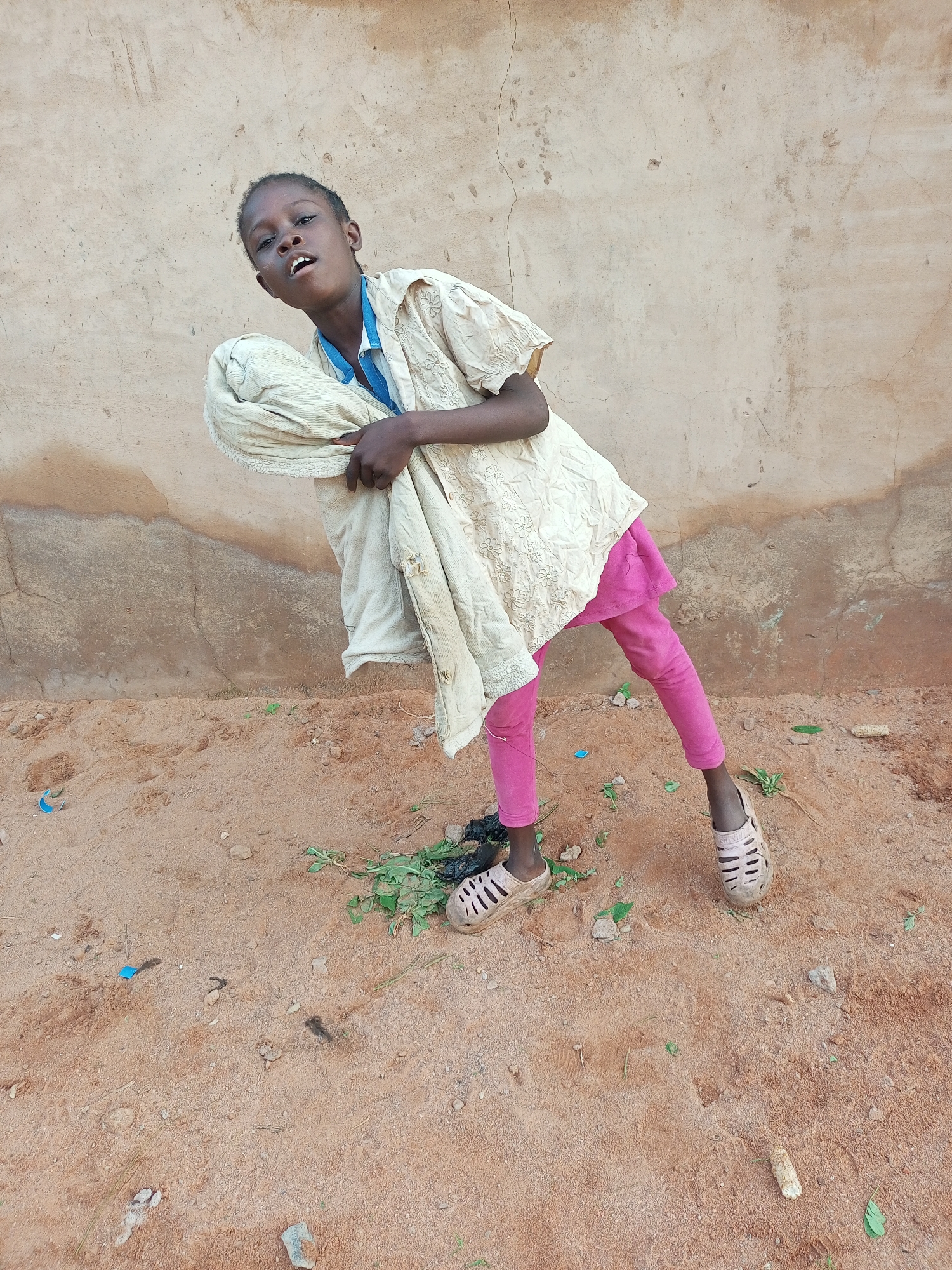 The image shows a young child standing in front of a textured, light-brown wall. The child is wearing a light-colored, textured shirt over a blue collar, and pink leggings. They are holding a light-colored fabric bundle. The ground is sandy and uneven, with scattered debris including a blue plastic object, green leaves, and a cob. The overall environment suggests an outdoor, possibly informal setting. The image was taken in Tempure, Nigeria.