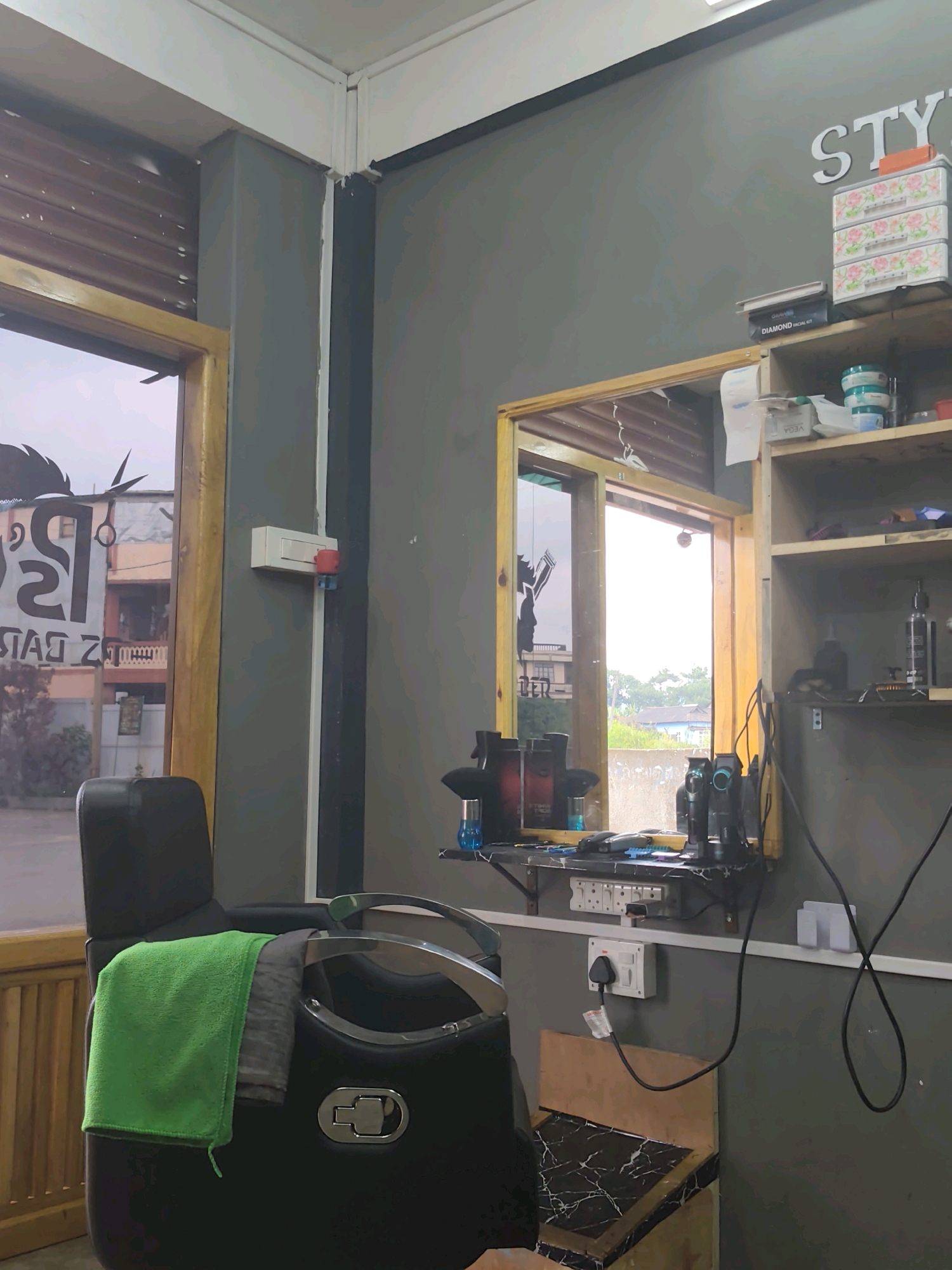 The image shows the interior of a barbershop, likely in Raliang, India. The foreground features a black barber chair with a green towel draped over its armrest and a grey garment folded on the seat. To the left of the chair is a window with wooden trim, revealing a glimpse of buildings and foliage outside. The barbershop walls are painted a dark gray. To the right, a mirror reflects the interior, showing a shelf above with various products, including containers and a "DIAMOND FACIAL KIT" box. Below the mirror is a shelf laden with hair clippers and grooming supplies. A power strip with multiple outlets is mounted on the wall, with one plug connected to a power source on the floor. A light switch is visible on the wall next to the window. The overall impression is of a functional and organized barbershop.