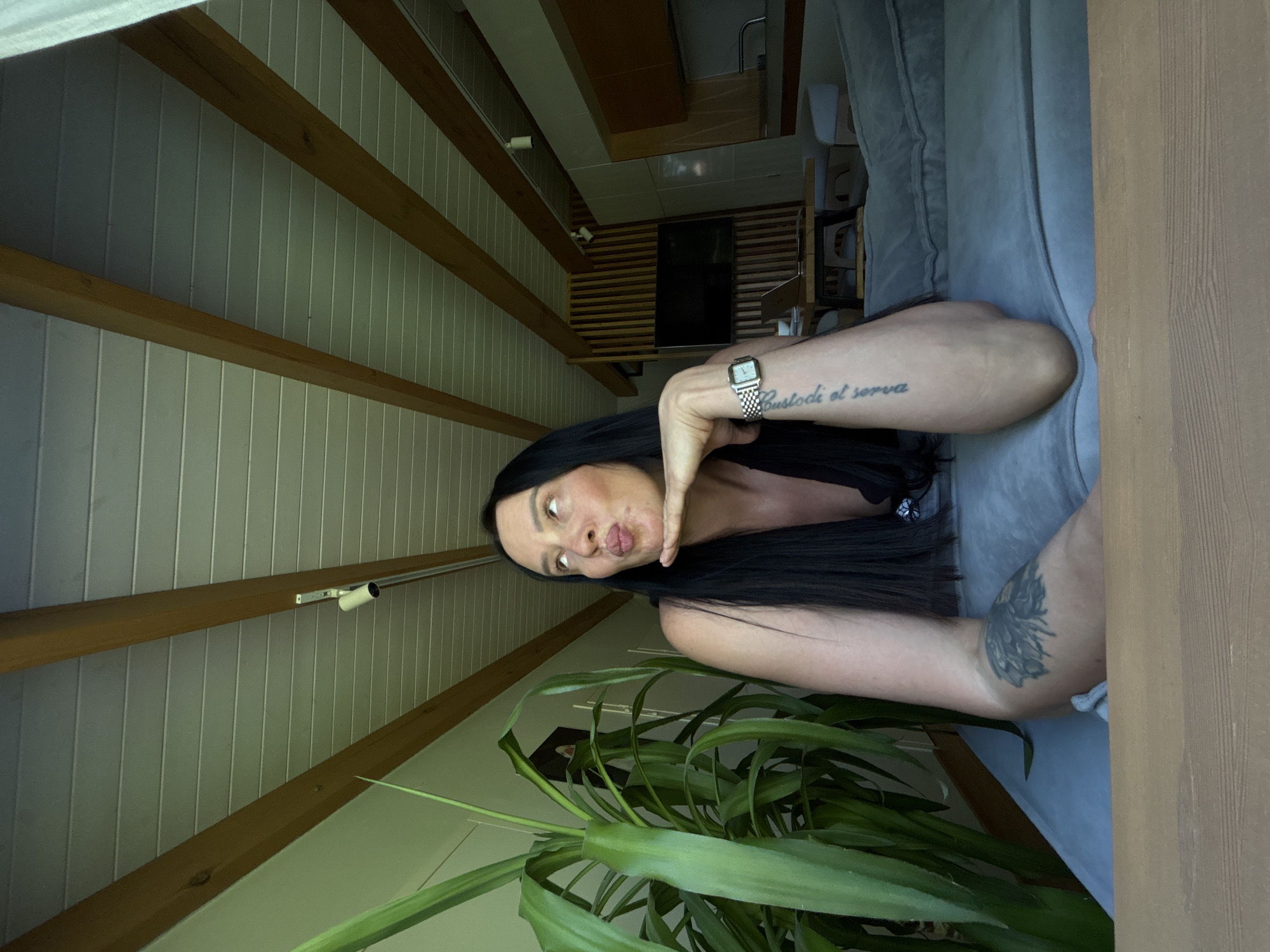 The image presents a woman with long, black hair, light skin, and tattoos on her arms. She is wearing a watch on her left wrist. She is holding her hand near her face in a posed manner. A large green plant is positioned in the foreground. The background reveals a room with a slanted ceiling featuring wooden beams and white paneling. A television and other furniture can be seen in the distant background. There is no indication that the photo was taken in the city of Chaikovsky, Russia.