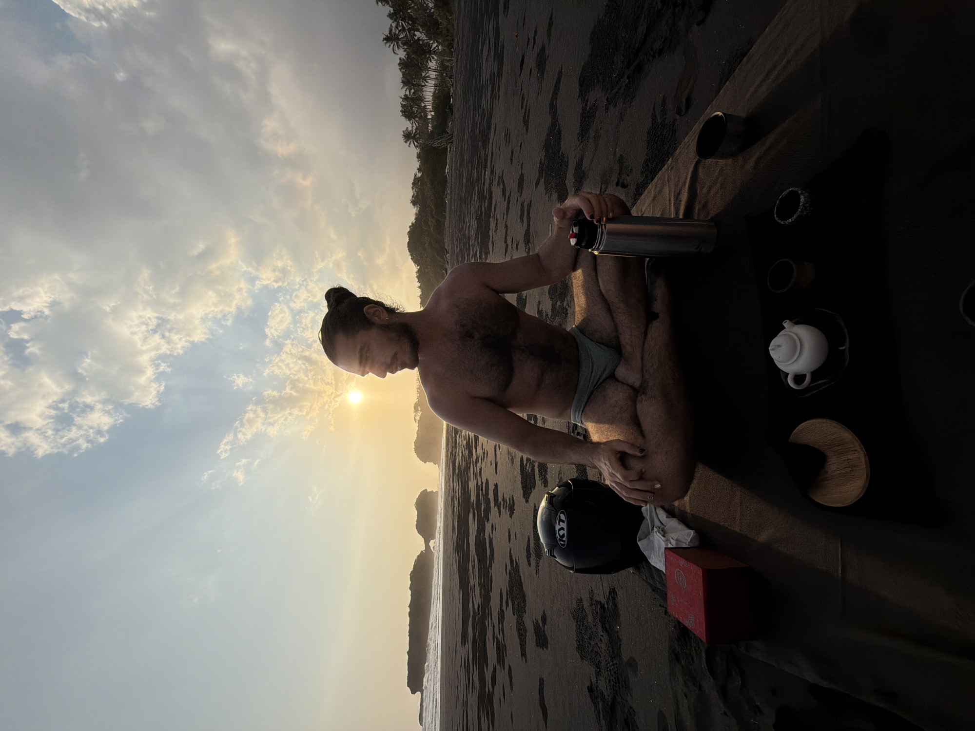 A man with a bun hairstyle and a beard is sitting in a lotus position on a dark mat on a black sand beach in Selemadeg, Indonesia. He is looking to his right, with the sun setting behind him, casting a warm glow. The man is shirtless and wearing light-colored swim trunks. In front of him on the mat, there is a thermos, a red box, a white teapot with two small cups, and a wooden coaster. A black motorcycle helmet with "KYT" branding is placed to his left, near the edge of the mat. The background shows the ocean with gentle waves and rocky outcrops in the distance, as well as palm trees lining the coast. The sky is filled with clouds, with sun rays breaking through.
