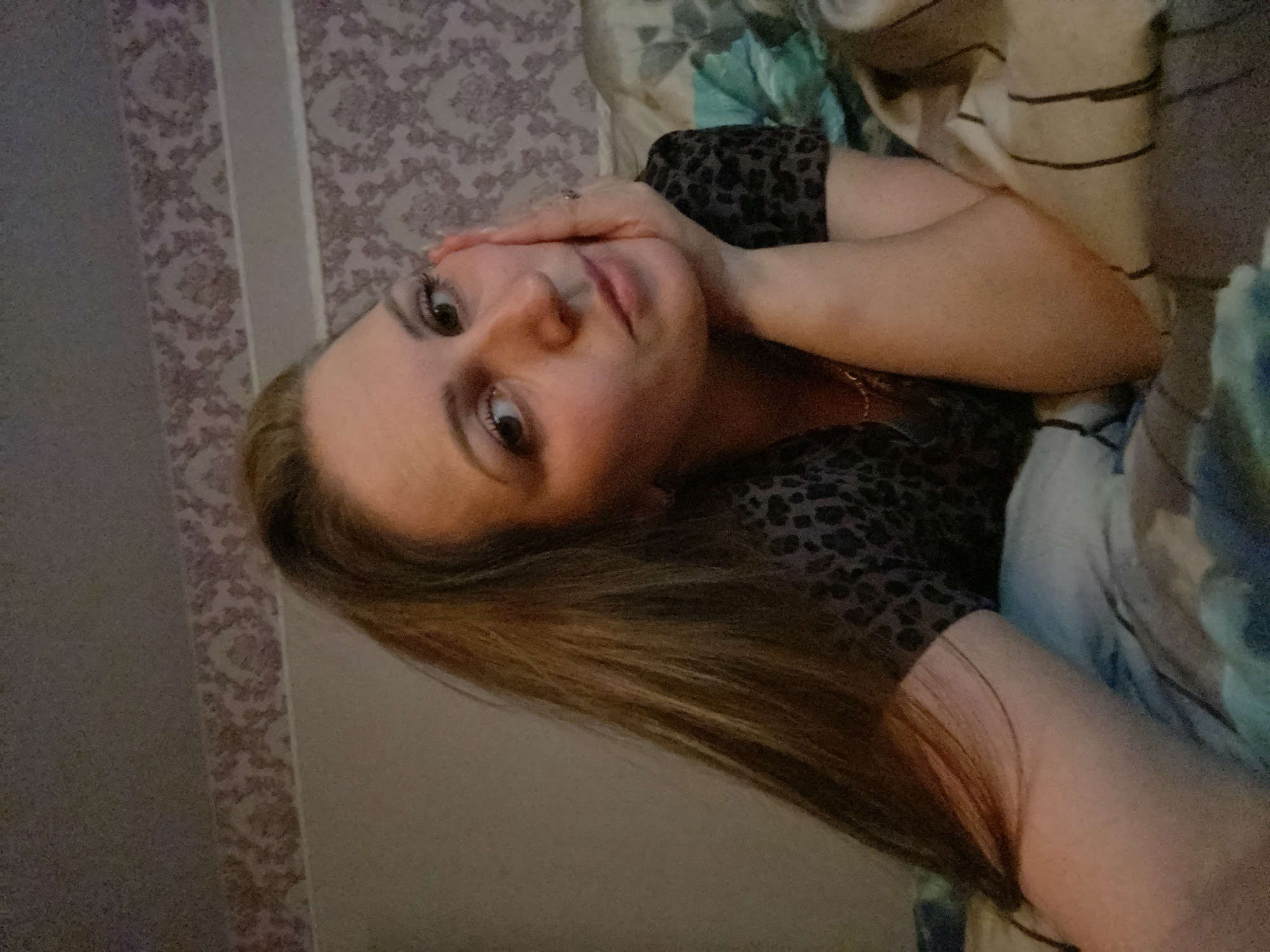 A woman is lying on her side on a bed. She is propped up on one arm, with her hand touching her forehead. She is wearing a leopard-print top. The background includes a patterned wall and bedding. The location is Markovskii, Russia.