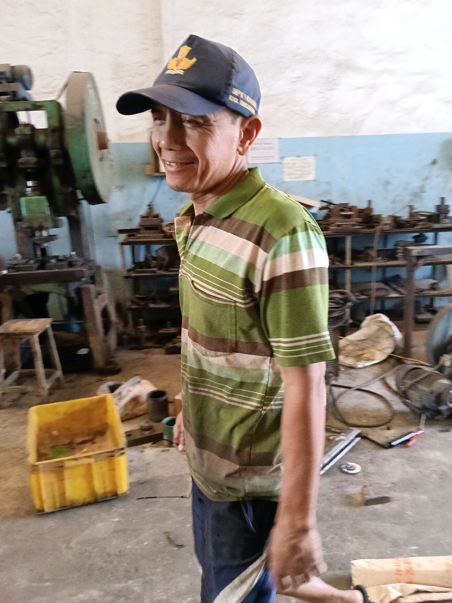 A man wearing a blue baseball cap and a green and brown striped short-sleeved polo shirt is standing in what appears to be a workshop or industrial space in Belimbing, Indonesia. He is looking to his left and smiling. In the background, there are various pieces of machinery, shelves filled with metal parts, and a yellow plastic bin on the floor. The environment appears to be concrete with some dirt and debris. The lighting suggests it is daytime.