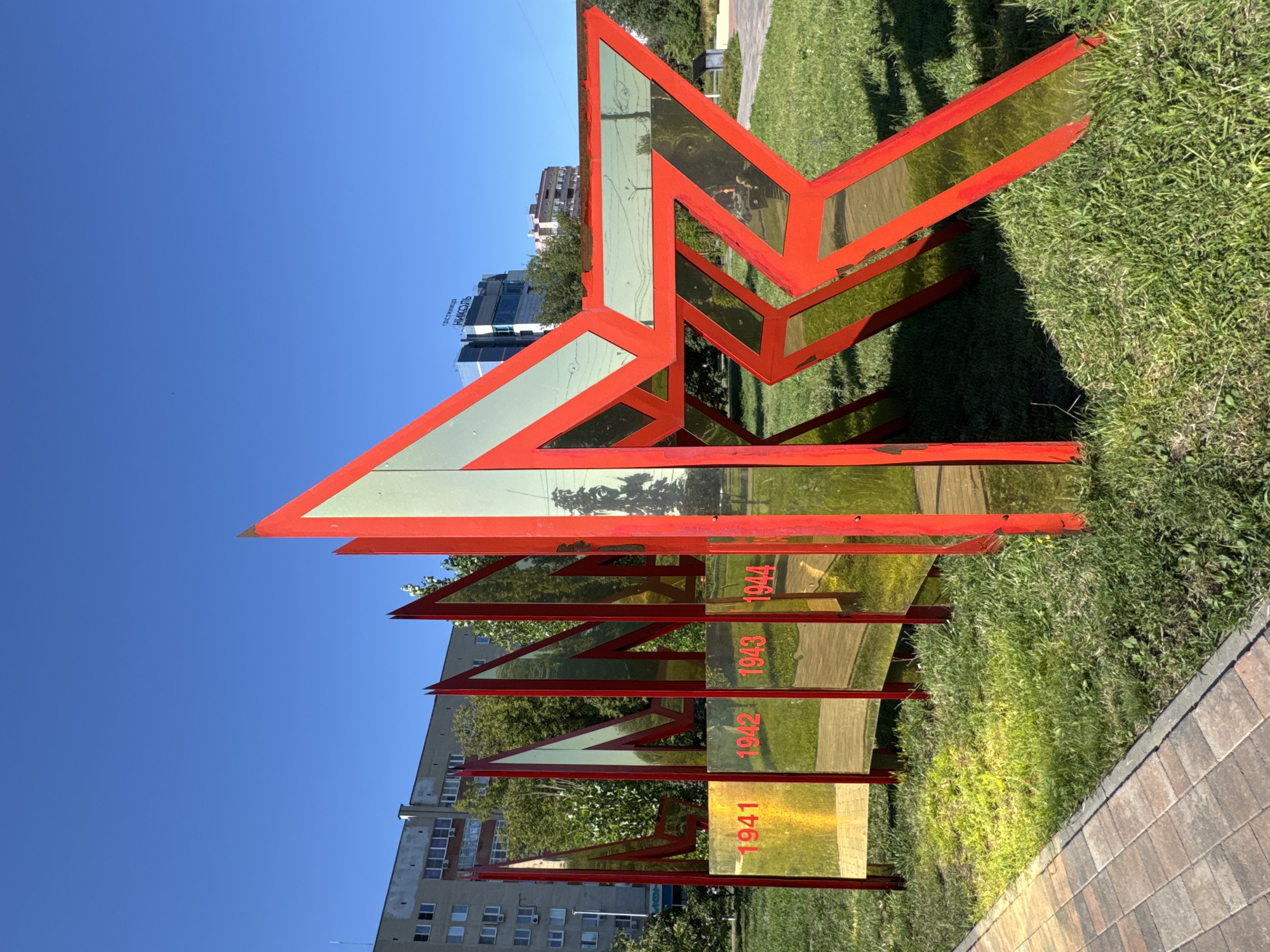The image shows a red star monument with reflective panels in a park in Nizhny Novgorod, Russia. The monument has red frames with what appear to be yellow or gold reflective panels. Each panel is shaped like a part of a star, and they are arranged in a staggered, almost zigzagging pattern. The front-most panels are inscribed with the years "1941", "1942", "1943", and "1944" in red lettering.
Behind the monument, there is a well-maintained lawn and a paved walkway. In the background, there are trees and several buildings. One building is a multi-story apartment complex with visible windows and balconies. Another building, taller and more modern, has "ГОСТИНИЦА НИКОЛЬ" (Hotel Nikoll) written on it. The sky is a clear, bright blue. The overall scene suggests a bright, sunny day. The monument appears to be a memorial, possibly related to a historical event like World War II given the dates inscribed on it.