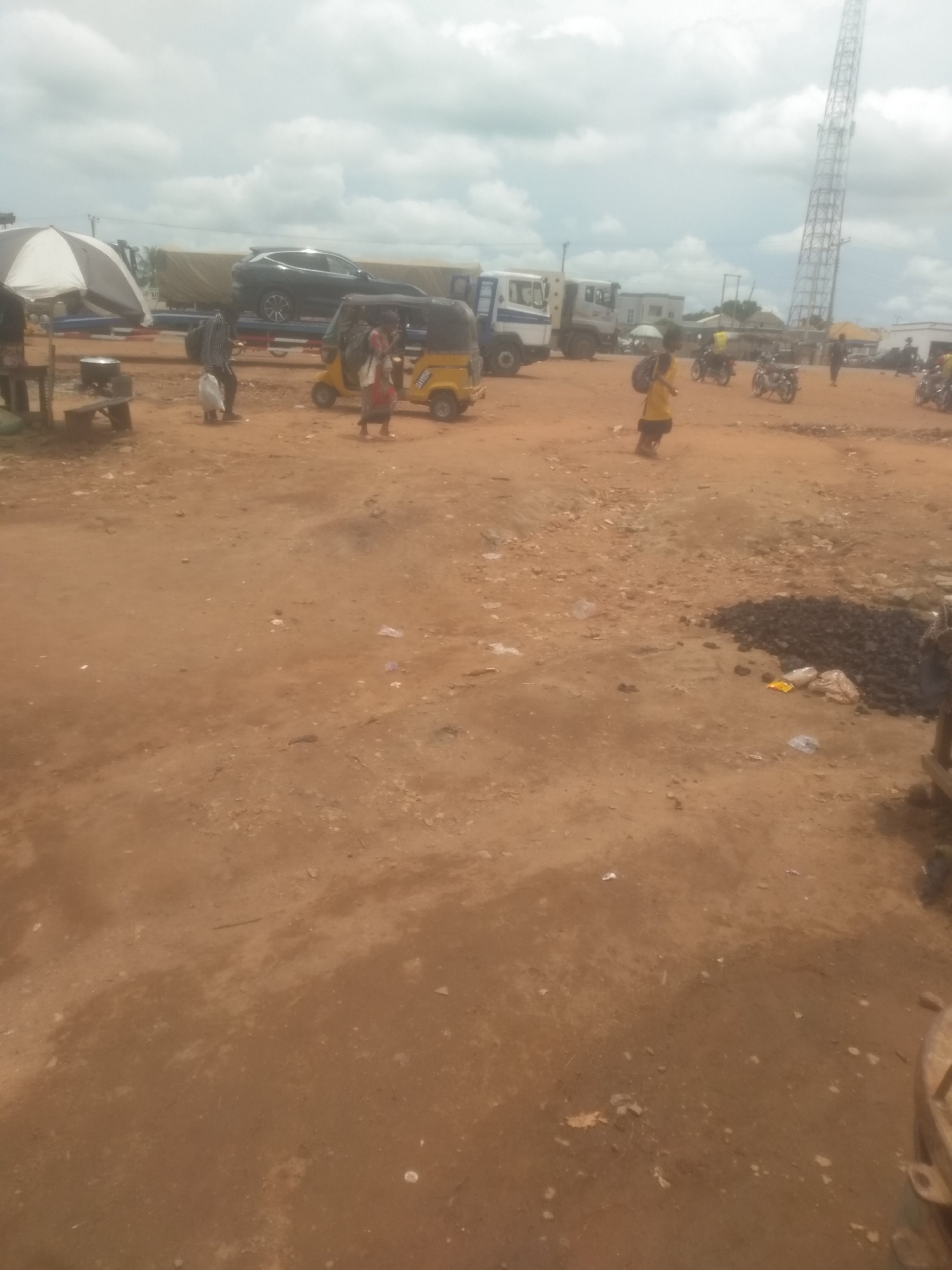 An outdoor, wide shot captures a busy market scene under a cloudy sky in an urban environment. The ground is composed of reddish-brown dirt, scattered with debris and small piles of dark, granular material. A prominent, tall metal tower with a lattice structure rises on the right side of the frame, piercing the overcast sky.
In the mid-ground, a yellow, three-wheeled auto-rickshaw is parked, with a woman in a colorful outfit standing nearby. To the left of the auto-rickshaw, a person in a dark shirt and pants walks while holding a white bag. Another individual, possibly a child, dressed in yellow and carrying a backpack, stands in the center of the frame.
Several vehicles are present in the scene. A black SUV is loaded onto a flatbed trailer, which is part of a larger transport truck. Other trucks and vehicles are visible in the background, some with their tarps covering their loads. Several motorcycles are also parked in the area.
On the far left, a white and grey umbrella is set up over a makeshift stall, with a metallic pot or container visible on a wooden bench. The overall impression is of a bustling marketplace with ongoing activity, likely a transportation hub or informal trade area.
