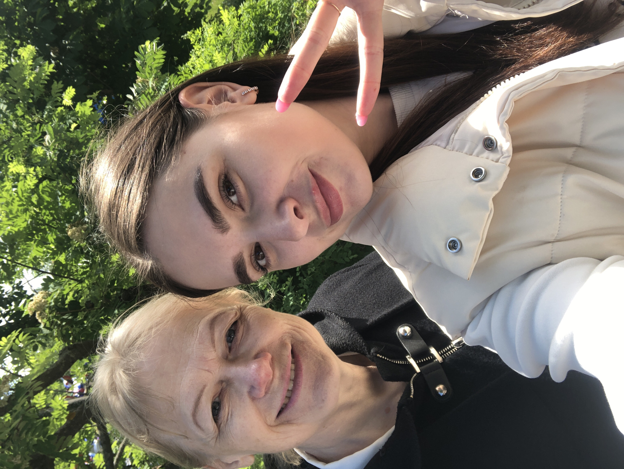 Two women are in the image. One is younger with long, dark brown hair, making a peace sign with her right hand. She is wearing a white jacket, presumably outside. The other woman is older with blonde hair. She is wearing a black jacket. Both women are smiling and appear to be taking a selfie. The background appears to be a park or garden with green foliage. The location is Chaikovsky, Russia.
