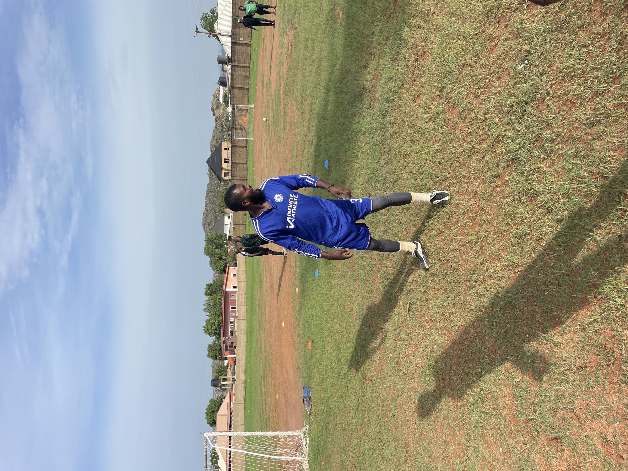 The image depicts a soccer player in a blue uniform stretching on a grassy field in Jos, Nigeria. The player is wearing black leggings under his shorts and has his shadow cast on the ground. The field is marked with orange and blue cones. In the background, there is a goalpost, a fence, buildings, and a rocky landscape. Two individuals in athletic wear stand in the distance. The sky is blue with some clouds.