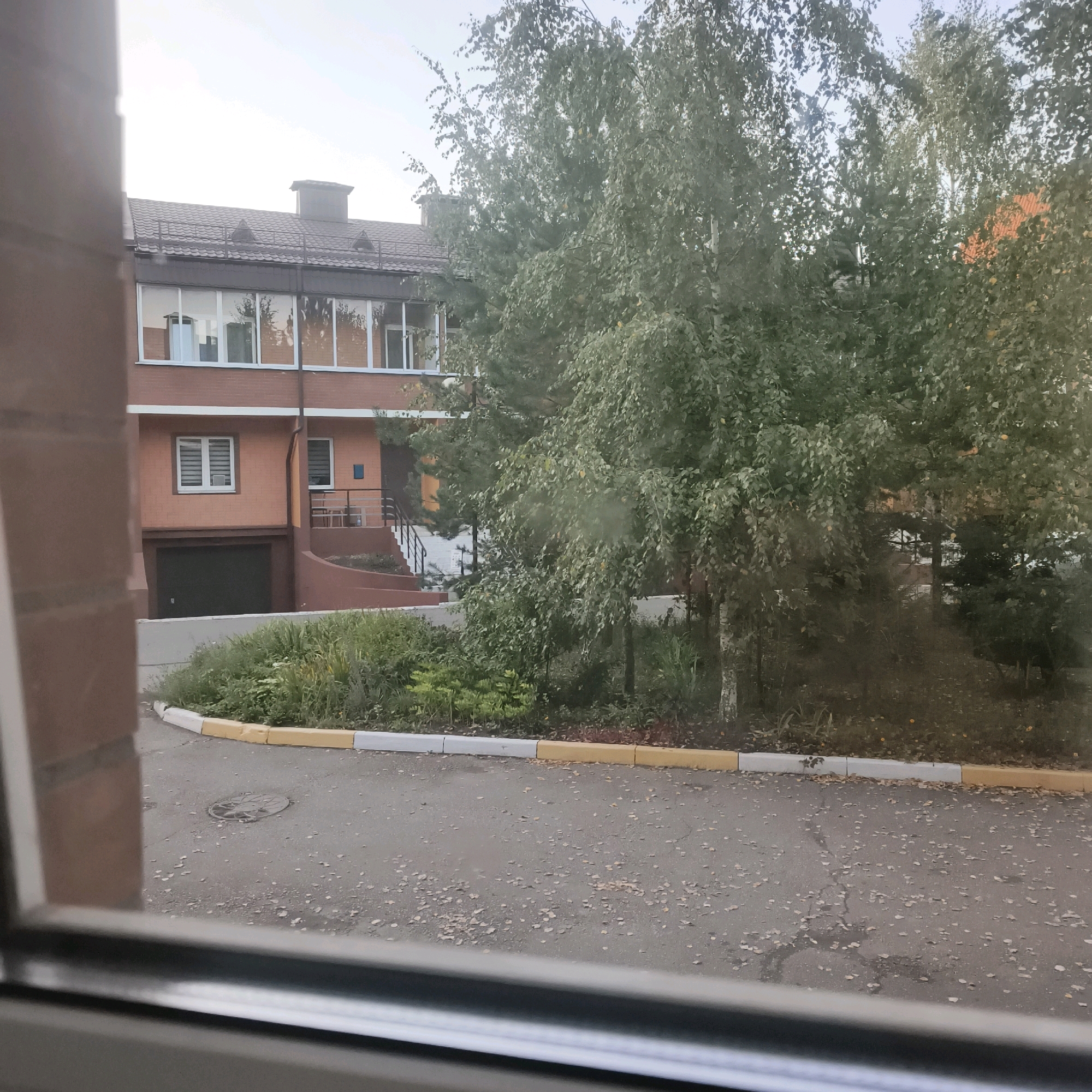 The image shows a view from a window, looking out onto a brick building with a dark tiled roof and large windows. In front of the building is a landscaped area with green bushes and trees, and a paved area with a manhole cover. The pavement is lined with a yellow and white curb. A brick wall is visible in the foreground, partially obscuring the view. The sky is overcast. The scene appears to be in a residential or commercial area. Based on the provided context, this scene is located in the city of Lunyovo, Russia.