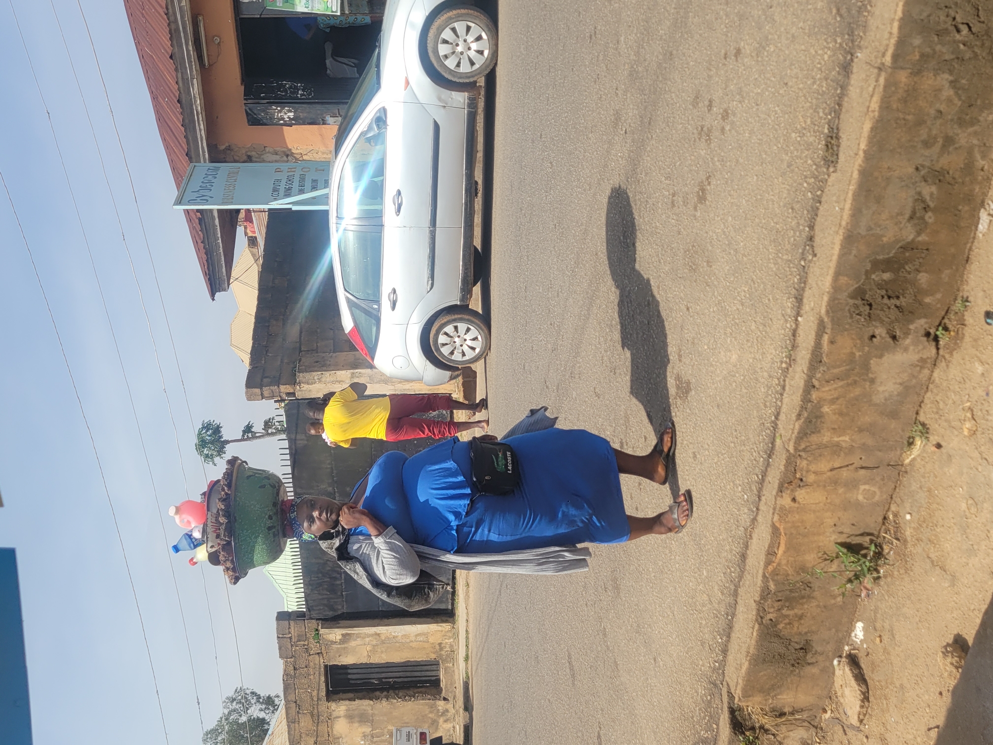 A woman in a blue dress and sandals walks along a paved street in Jos, Nigeria. She carries a large, green, bowl-like container on her head, which holds various items including plastic bottles and a pink object. A grey shawl is draped over her shoulder, and she wears a black "Lacoste" brand shoulder bag. A person in a yellow shirt and red pants is walking in the background. A white car is parked beside a building with a sign that says "Cybercom Business Central". The photograph is taken on a sunny day.