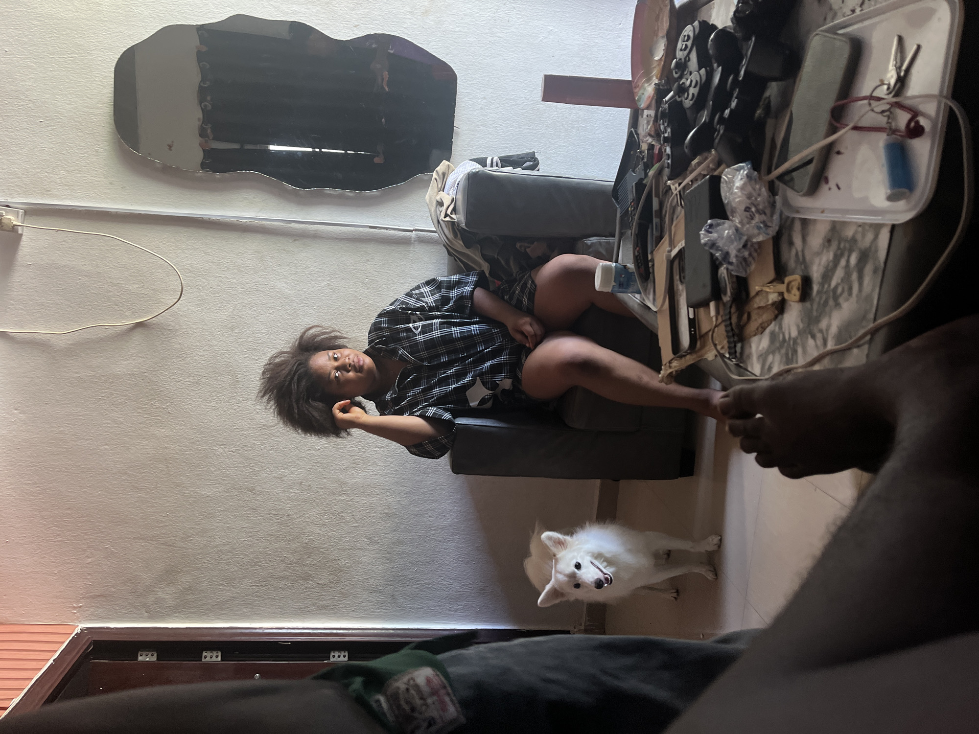 An interior scene featuring a woman seated on a gray armchair positioned against a white wall, with an irregularly shaped mirror above. The woman has dark hair and wears a patterned black and white shirt. A white dog stands near her feet on a tile floor. Adjacent to the armchair is a table covered with objects including electronic devices, controllers, and small containers. The presence of an individual's legs in the foreground suggests another person is taking the picture. The location is specified as Ikot Ekpene, Nigeria.