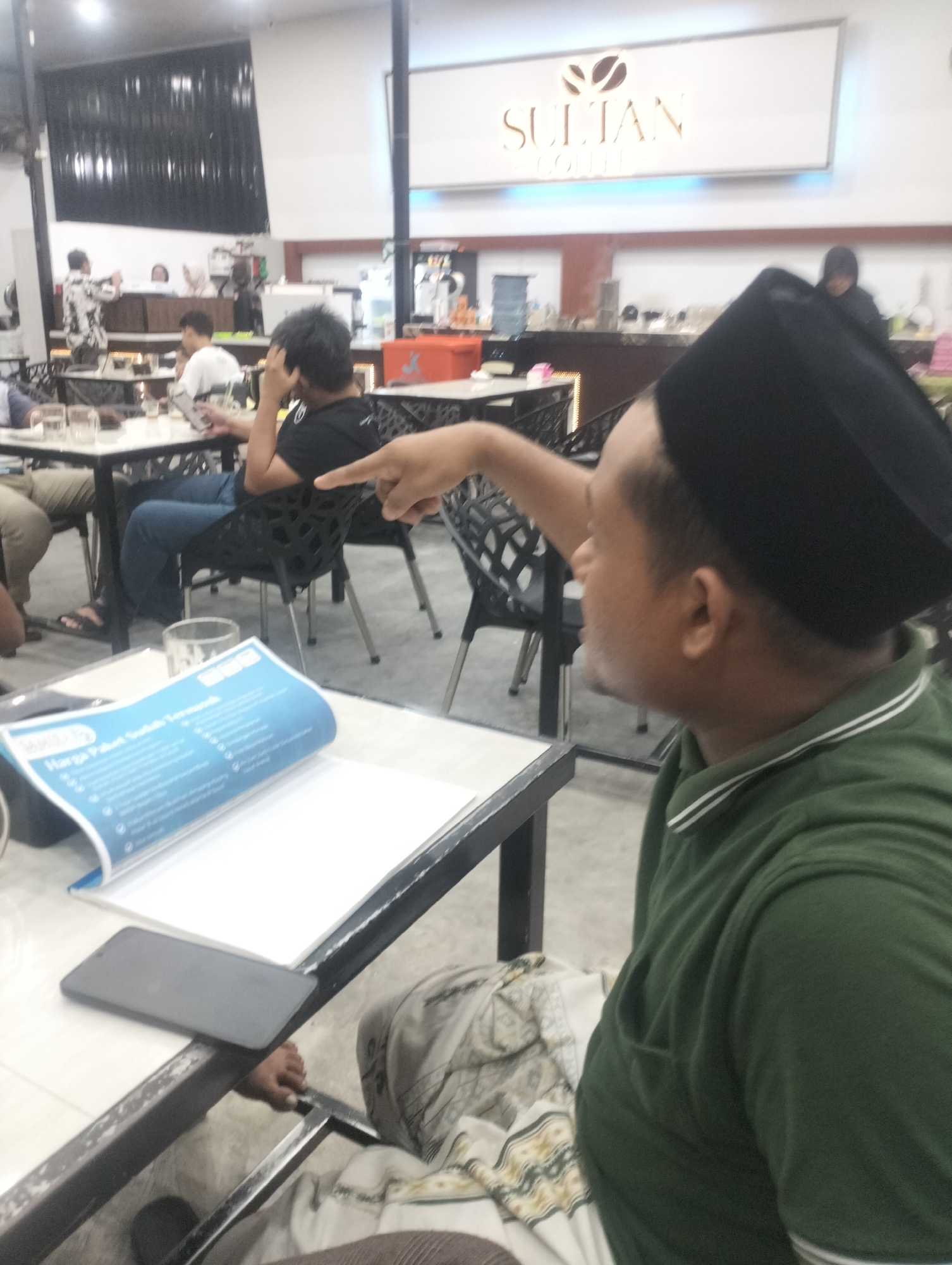 In a coffee shop setting, likely in Lhokseumawe, Indonesia, a man wearing a black peci hat and a green polo shirt with white trim is captured in profile, pointing towards the left of the frame. His gaze is directed off-camera. The background reveals other patrons seated at tables, some with their backs to the viewer. One individual in the mid-ground, wearing a black t-shirt, is looking at a smartphone. Another person in a white shirt is also seated at a table, appearing to be engaged with their phone.
In the foreground, a white table holds a smartphone lying flat and a blue brochure or menu, partially open. The brochure displays text in a foreign language, with the heading "Harga Paket Sudah Termasuk" visible. This translates to "Package Price Already Includes" in Indonesian, suggesting a menu or service offering. The overall lighting suggests an indoor setting, possibly during the day or evening, with artificial lights illuminating the space. The ambiance appears casual and relaxed, typical of a coffee shop. The logo for "SULTAN COFFEE" is visible on a sign in the background. The people in the scene are casually dressed, indicating a relaxed social gathering or a break at a coffee shop.