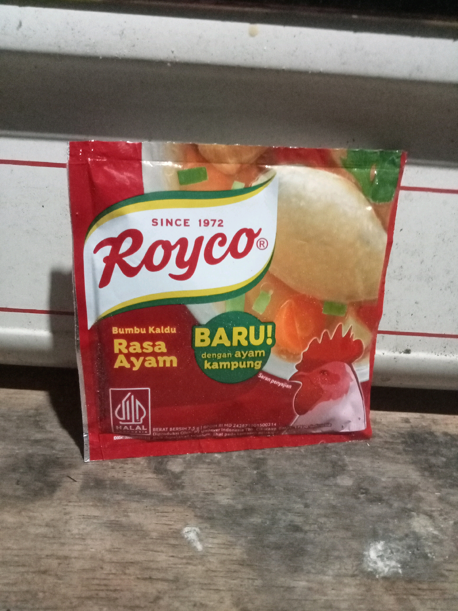 The image shows a red and white pouch of Royco Chicken Broth Seasoning, a food product from Indonesia, positioned on a wooden surface. The pouch has prominent branding with the Royco logo in red and white script, and text indicating "SINCE 1972," "Bumbu Kaldu Rasa Ayam," and "BARU!" The pouch also features an illustration of a rooster and vegetables. A "HALAL" logo is visible in the bottom left corner. The wooden surface appears to be a table or countertop, with some white powdery residue scattered on it. The background is out of focus, showing a textured, light-colored surface that might be a wall or shelf. There are no people or animals present in the image. The location is stated to be Rambeanak, Indonesia.