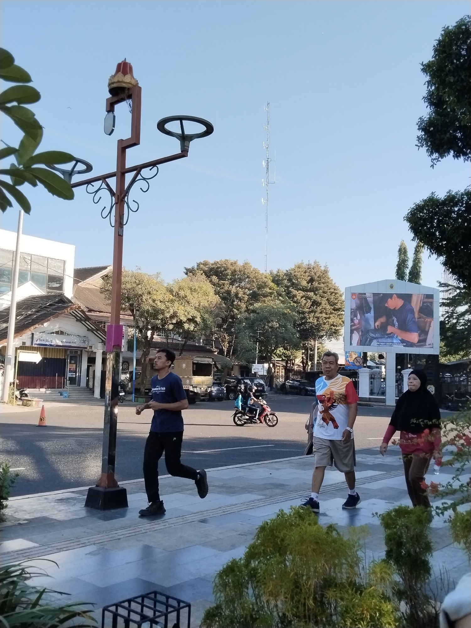 In the city of Kecamatan Pati, Indonesia, on a clear, sunny day, a street scene unfolds. A man in a dark blue t-shirt and black pants is jogging on a paved walkway. To his right, a man in a white t-shirt with a colorful graphic and khaki shorts walks, followed by a woman wearing a black hijab and a patterned red and pink outfit. They are walking on the same paved walkway. In the background, a large outdoor screen displays an image of a man. Across the street, a bank with the sign "Bank Jateng" is visible, alongside other buildings and trees. A street light with a decorative finial stands prominently on the walkway. Traffic, including motorcycles and cars, moves along the road. The overall environment suggests a typical urban setting during daylight hours.