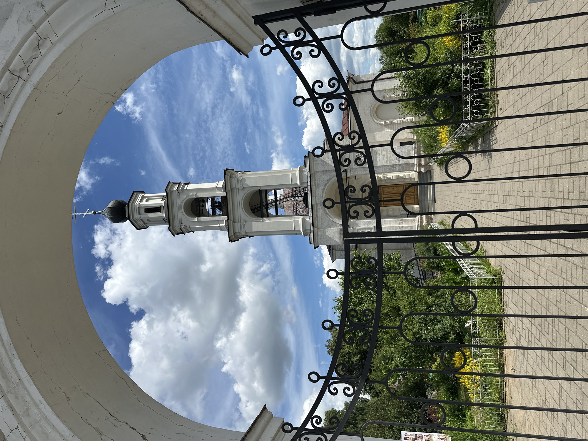 The image captures a view through a large, weathered arched gateway towards a white bell tower and a church in the distance. The gateway is made of white, cracked plaster. A black wrought-iron gate, decorated with ornate scrollwork and circles, is partially open in the foreground, obscuring a paved walkway leading to the church.
The bell tower is tall and slender, painted white, with multiple tiers and arched openings. It is topped with a dark dome and a cross. The church itself is also white, with classical architectural details, including columns and arched windows. A red-tiled roof is visible behind the bell tower.
The area in front of the church appears to be a garden with lush green trees and bushes. Patches of bright yellow flowers are visible amongst the foliage. The sky above is a vibrant blue, dotted with fluffy white clouds. The scene is bathed in bright daylight, suggesting a sunny day.
The location is identified as the Church of the Resurrection in the village of Yerynino, Russia. There are no people visible in the image.