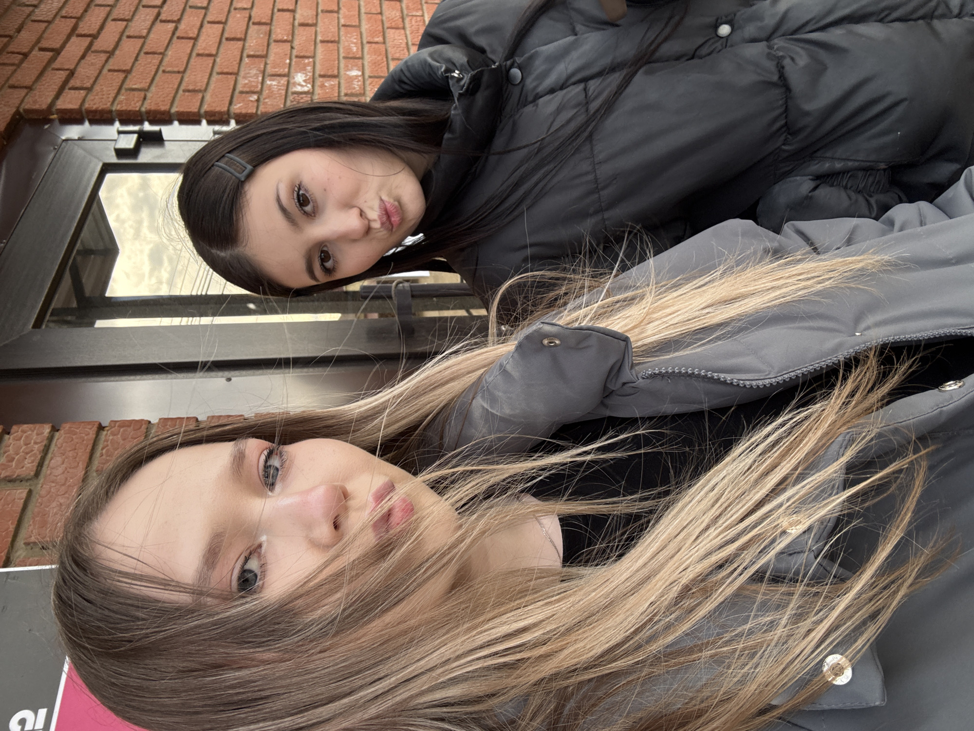 This media file captures two young women posing for a casual photo outdoors, presented with a 90-degree clockwise rotation. The setting is in Фоки, Russia, during what appears to be daytime, indicated by the natural light. Both individuals are dressed in warm puffer jackets, suggesting cool weather.
In the foreground, a woman with long, light blonde and brown hair partially covering her face looks towards the camera. She has light-colored eyes and fair skin and is wearing a grey puffer jacket with visible metallic snaps.
Behind her, another woman with long dark hair and dark eyes is visible, making a playful "duck face" with puckered lips. A dark hair clip is secured in her hair. She is wearing a black puffer jacket, with details like a button and textured fabric visible.
The background shows a building with a red brick facade and a dark-framed window or door, suggesting an urban or suburban environment. On the far left edge, part of a sign with pink and black colors and the partial text "И!" is visible. The overall mood of the image is informal and friendly.