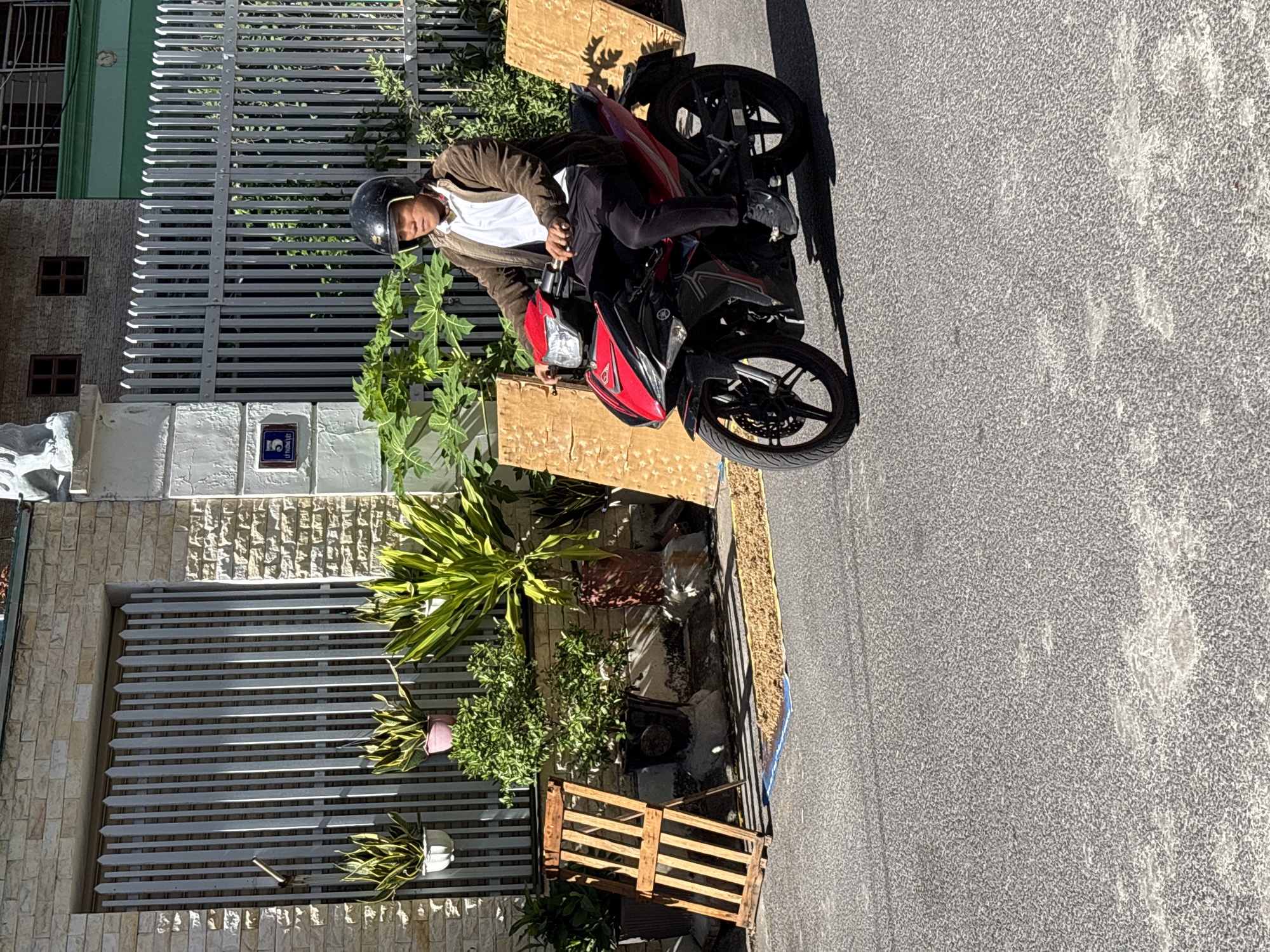 A man wearing a helmet, brown jacket, and white shirt rides a red and black motorcycle on a paved street. He is in motion, looking forward. The background shows a residential area with a stone wall, a metal gate, and lush green plants. There are also two large wooden panels propped up on the side of the road. A small patch of brown material, possibly dried goods, is visible on the ground near the wooden panels. The scene appears to be in Nha Trang, Vietnam, as indicated by the street sign with Vietnamese text.