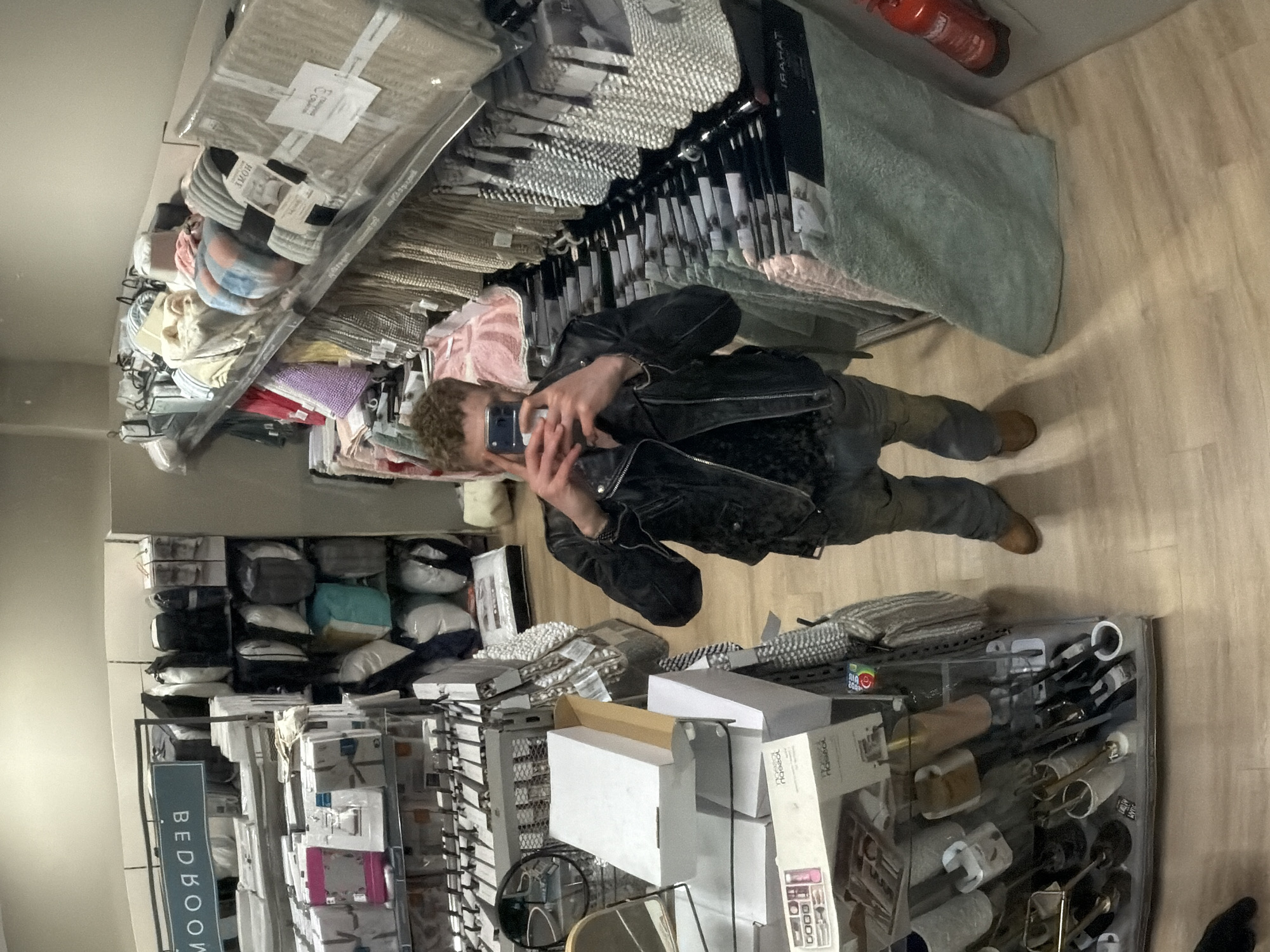 The image shows a person taking a selfie, likely reflected in a mirror or glass surface, inside a retail store. The subject has light, curly hair and is wearing a black leather jacket, dark trousers, and light brown boots. They are holding a dark-colored smartphone to capture the image.
The setting is an indoor retail environment, possibly a home goods or department store, indicated by shelves filled with various products. To the left and behind the person are multiple shelves stacked with packaged textiles, including bedding, pillows, and towels in various colors and patterns. A prominent sign on a display rack clearly reads "BEDROOM."
Other notable objects include a display case in the foreground on the right, containing an assortment of smaller items such as decorative objects, boxes, and what appear to be household accessories. A red fire extinguisher is visible mounted on the wall in the upper right corner of the frame. The floor is made of light-colored wood planks. Visible text on product packaging includes "HOME," "ARAHAT" (reversed), "GUM DROP," and "Naoeao" (reversed). The lighting suggests an indoor, artificially lit environment.
