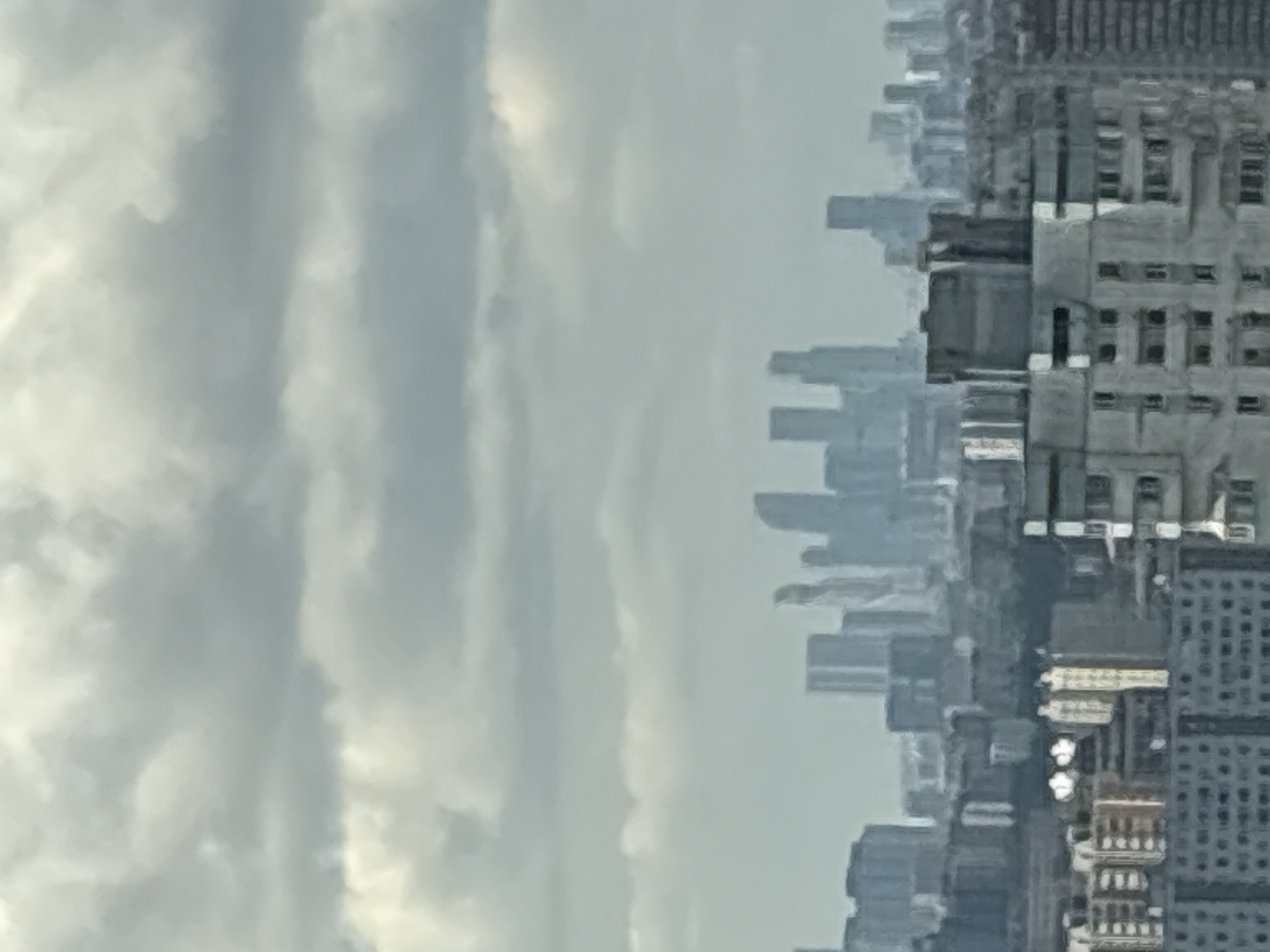 The image shows a cityscape with tall buildings under a cloudy sky. The buildings are densely packed and appear to be of varying heights and architectural styles. The sky is overcast with diffuse light, suggesting a hazy or foggy atmosphere. The perspective is somewhat elevated, as if the photograph was taken from a high vantage point.