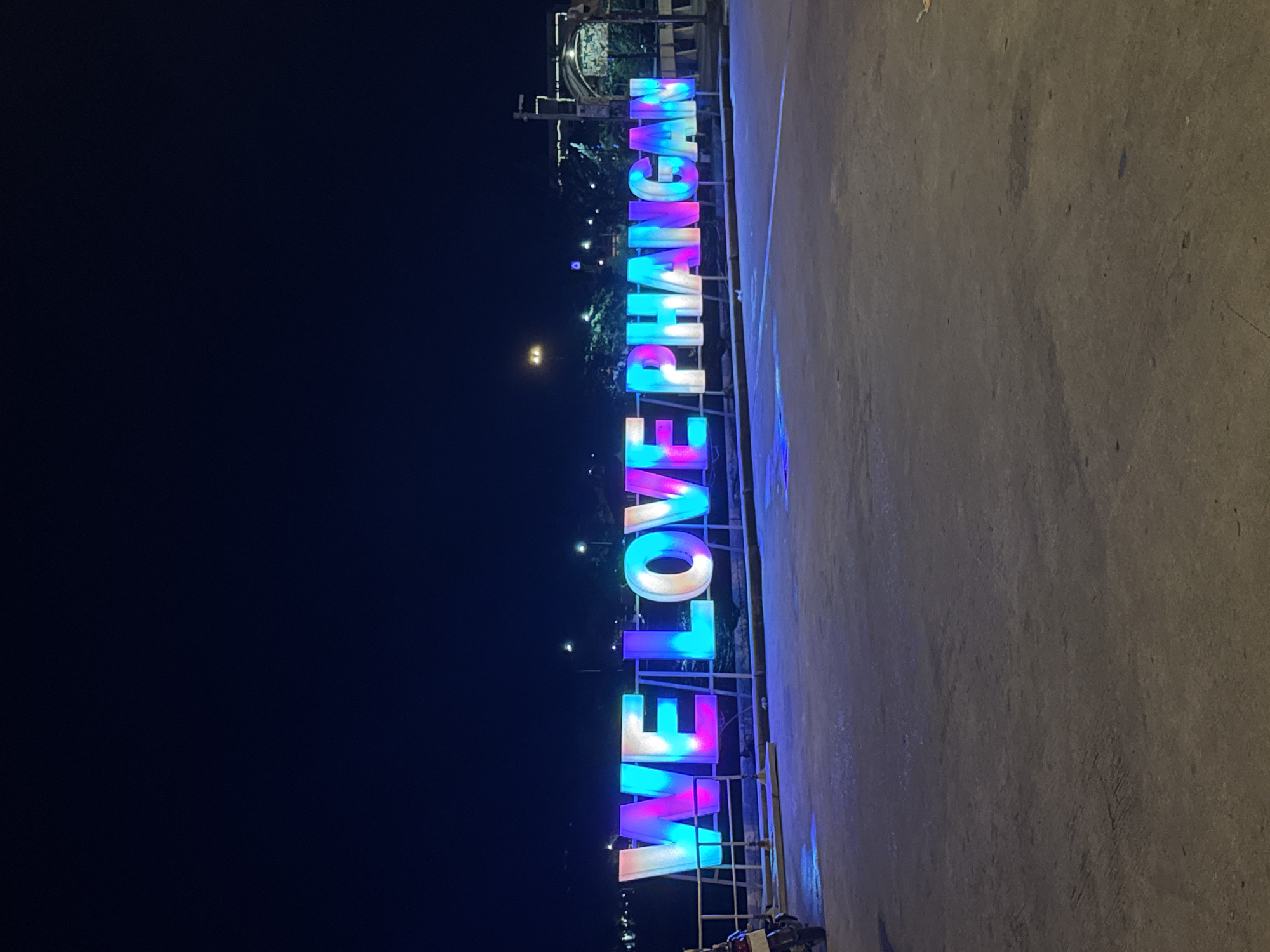 The image displays a nighttime scene in ตำบลเกาะพะงัน, Thailand, featuring a large, illuminated sign spelling "WE LOVE PHANGAN" in capital letters. The sign's letters exhibit dynamic color changes, predominantly showcasing shades of blue, pink, white, and purple. It is positioned on a raised platform or low barrier, with metal railings visible behind it.
In the foreground, a dark, textured paved surface, likely concrete or asphalt, extends across the bottom portion of the frame. The left edge of the frame shows a partial view of a dark-colored motorcycle.
The background is dark, indicating night, with numerous distant artificial lights scattered across what appears to be a shoreline or urban area. Several visible streetlights illuminate portions of the background. No people are discernible within the frame. The overall lighting is dominated by the sign's luminescence and the scattered background lights.