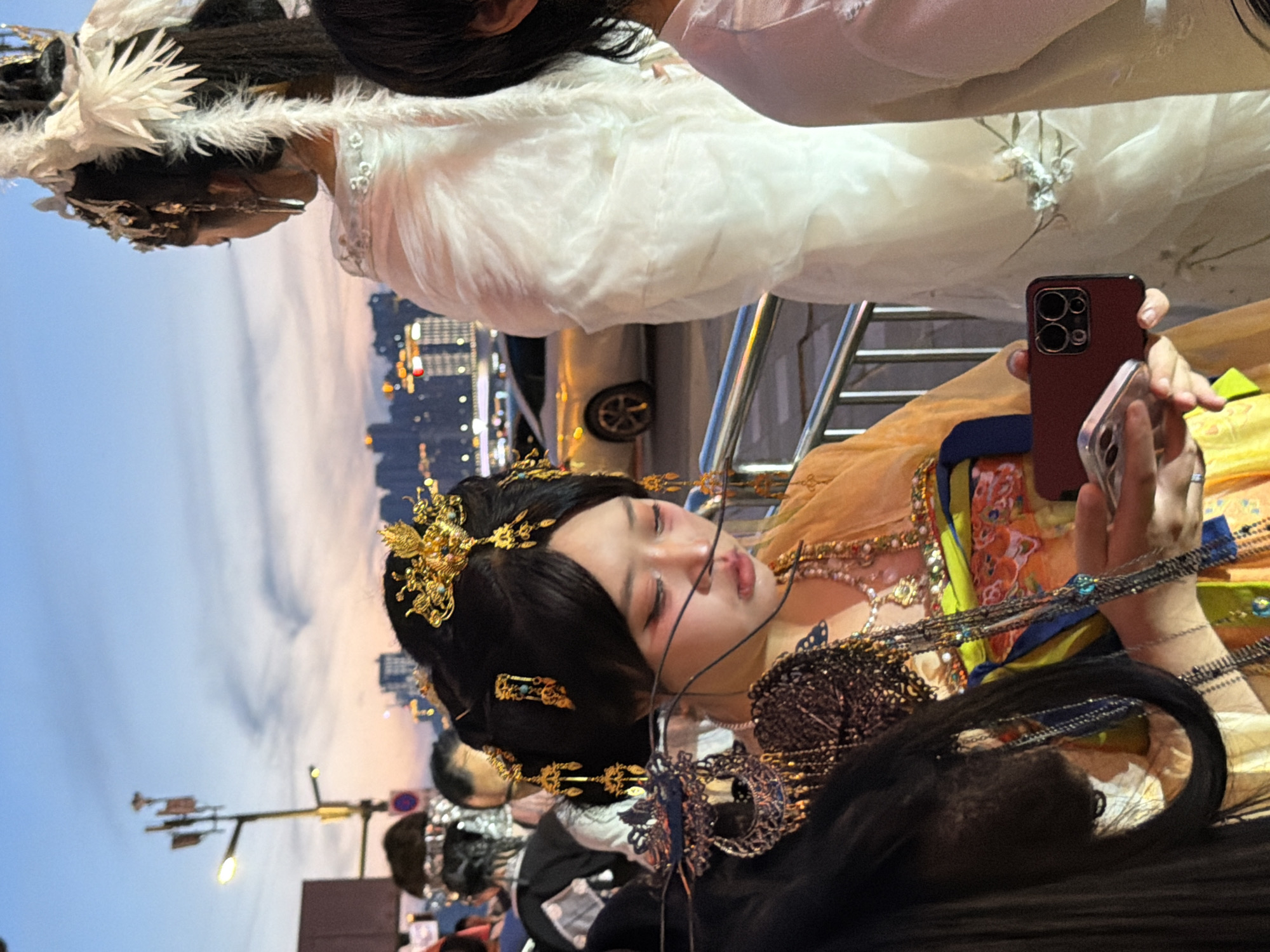 This is a close-up, eye-level shot of two women in traditional Chinese attire, with a city skyline in the background during twilight. The woman in the foreground, with black hair styled in an elaborate updo adorned with gold ornaments, wears an ornate yellow and orange garment. She is holding a dark red smartphone in her right hand and looking down at it. Her makeup includes dramatic red accents around her eyes. Multiple layers of beaded necklaces adorn her neck and chest.
To her right, another woman stands facing away, wearing a voluminous white layered garment that trails behind her. She also wears an elaborate headdress with white feathers. The background shows the Yuzhong District in Chongqing, China, with illuminated buildings and a road with a silver car visible. A metal railing separates the foreground figures from the street. The sky is a gradient of blue and orange, indicative of dusk. The overall scene captures a cultural or festive event.