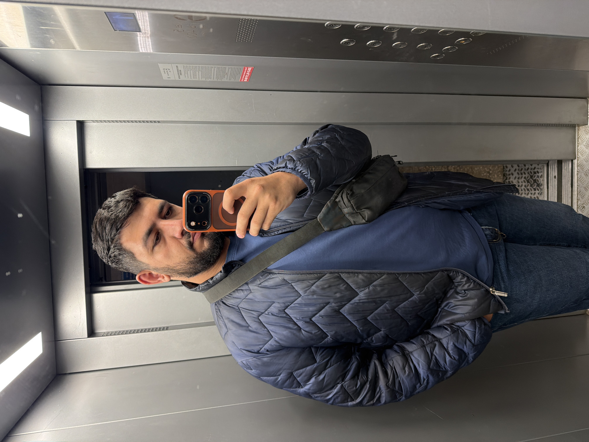 A man with short dark hair and a beard is taking a selfie in an elevator. He is wearing a blue quilted jacket, a blue t-shirt, and jeans. He is holding a smartphone with an orange case. He is also wearing a black bag over his shoulder. The elevator has a stainless steel interior with lights on the ceiling and buttons on the wall. The location cannot be determined from the image.