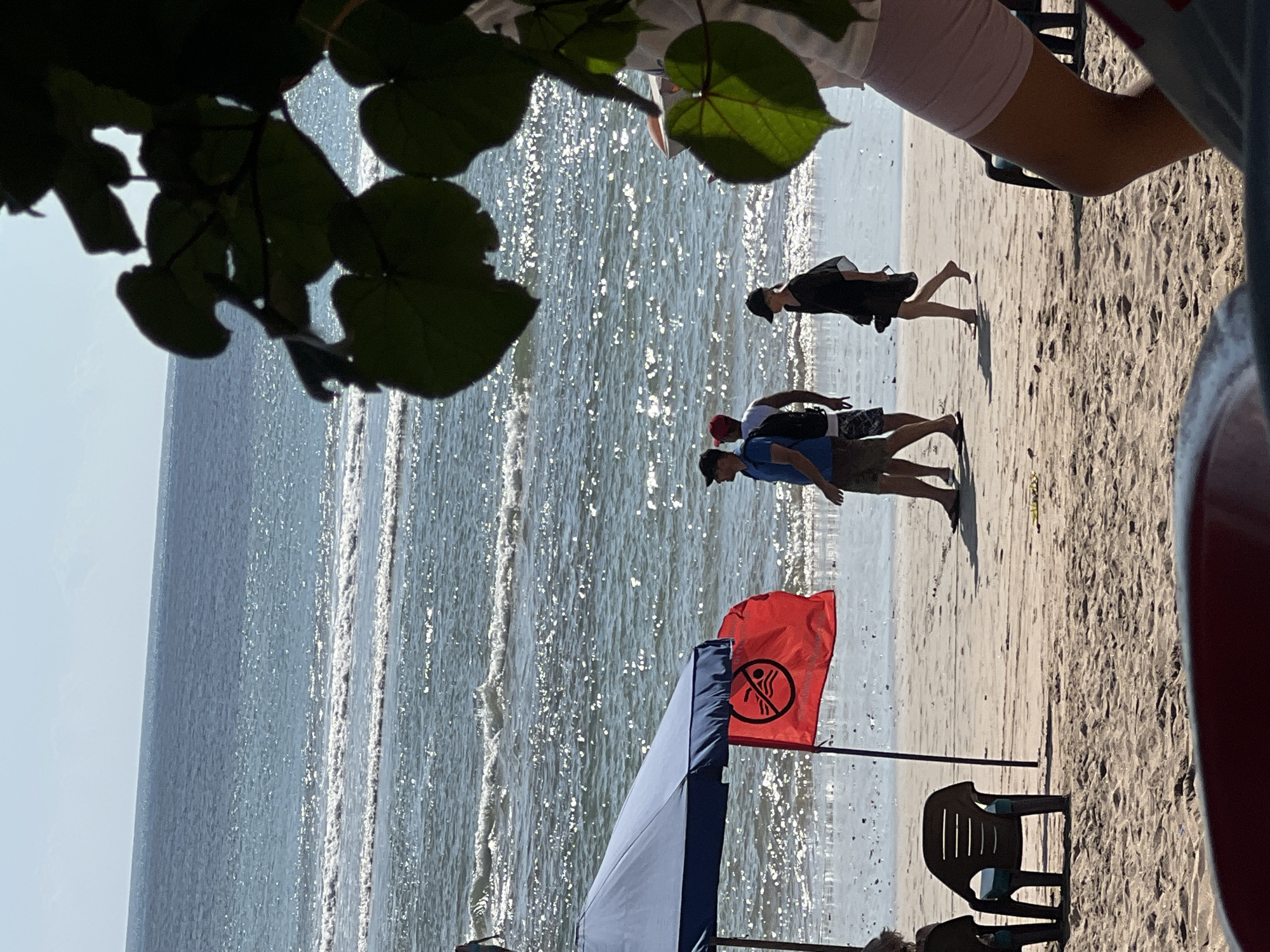 The image shows a beach scene in Kuta, Indonesia. In the foreground, part of a red picnic table is visible. To the left, a blue beach umbrella casts a shadow over two empty plastic chairs. A red flag with a black symbol of a crossed-out swimmer is planted in the sand next to the umbrella. In the middle ground, three individuals are walking on the wet sand near the water's edge. One person, wearing a blue t-shirt and dark shorts, walks with a black backpack. Another person in a white t-shirt and dark shorts is beside them. A third person in a black dress and a black hat is walking slightly ahead. The ocean is in the background, with gentle waves breaking on the shore and the sun glinting off the water. The sky is clear and bright. Lush green leaves are silhouetted in the upper right corner of the frame.