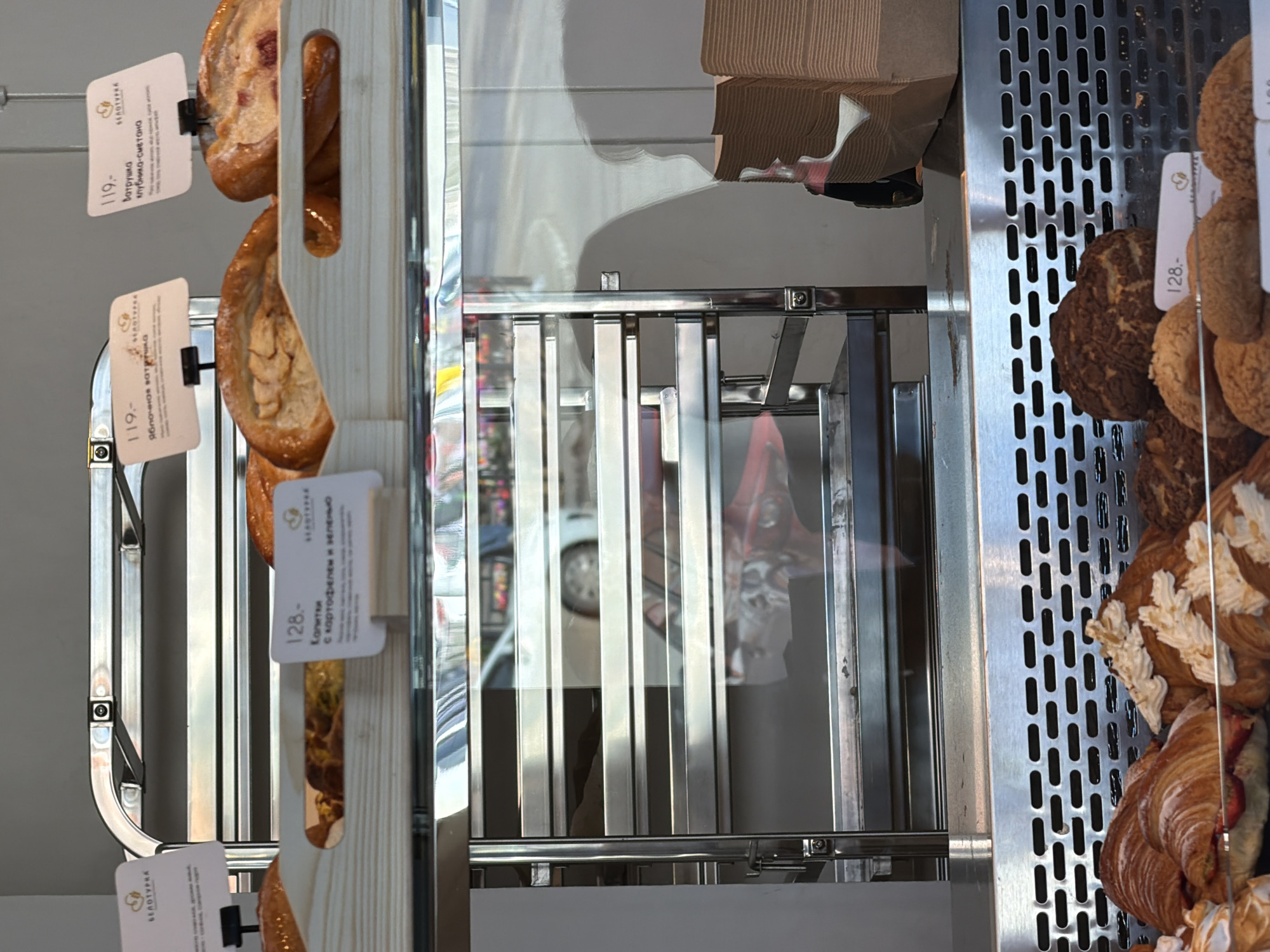 This indoor shot features a bakery display case filled with pastries and their associated price tags. On the left side of the frame, wooden stands hold pastries labeled with prices like "119" and "128." The price tags are written in Russian and seem to indicate the names of the pastries. On the right side, a perforated metal wall separates another section of pastries. These include croissants and other sweet treats. The reflection in the glass showcases an outdoor scene with a light-colored car and various reflections, adding depth to the image. The pastries appear to be freshly baked and are presented in an organized, appealing manner. Based on the signs in the image, the location is likely in Izhevsk, Russia.