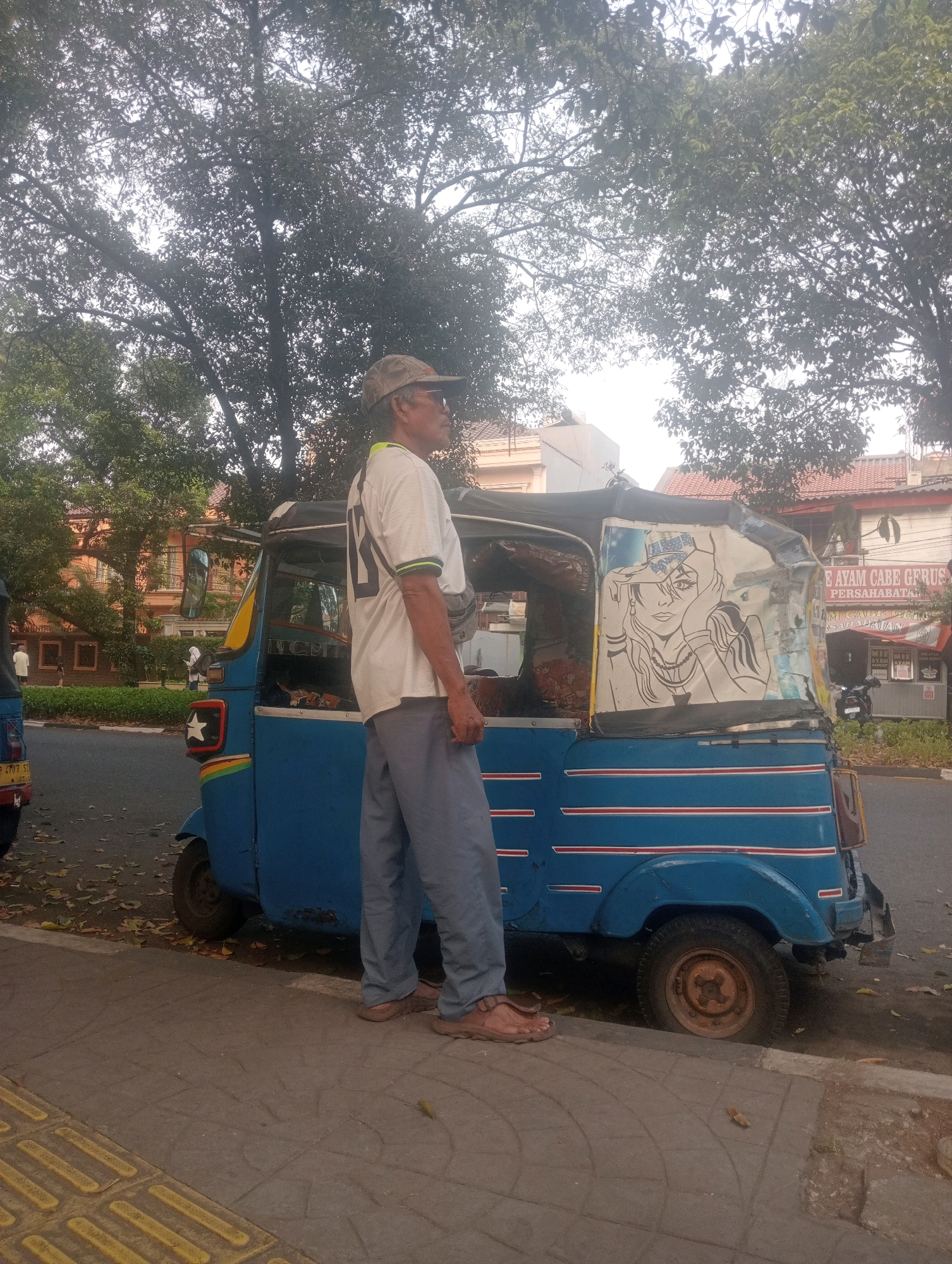 In East Jakarta, Indonesia, a man stands on a sidewalk next to a blue auto-rickshaw. The man, wearing a cap, sunglasses, a white t-shirt with black trim, and grey pants, is facing right. The auto-rickshaw, which has a drawing of a woman on its back, is parked on the side of a street lined with trees. Buildings with red tile roofs are visible in the background. Fallen leaves are scattered on the ground.