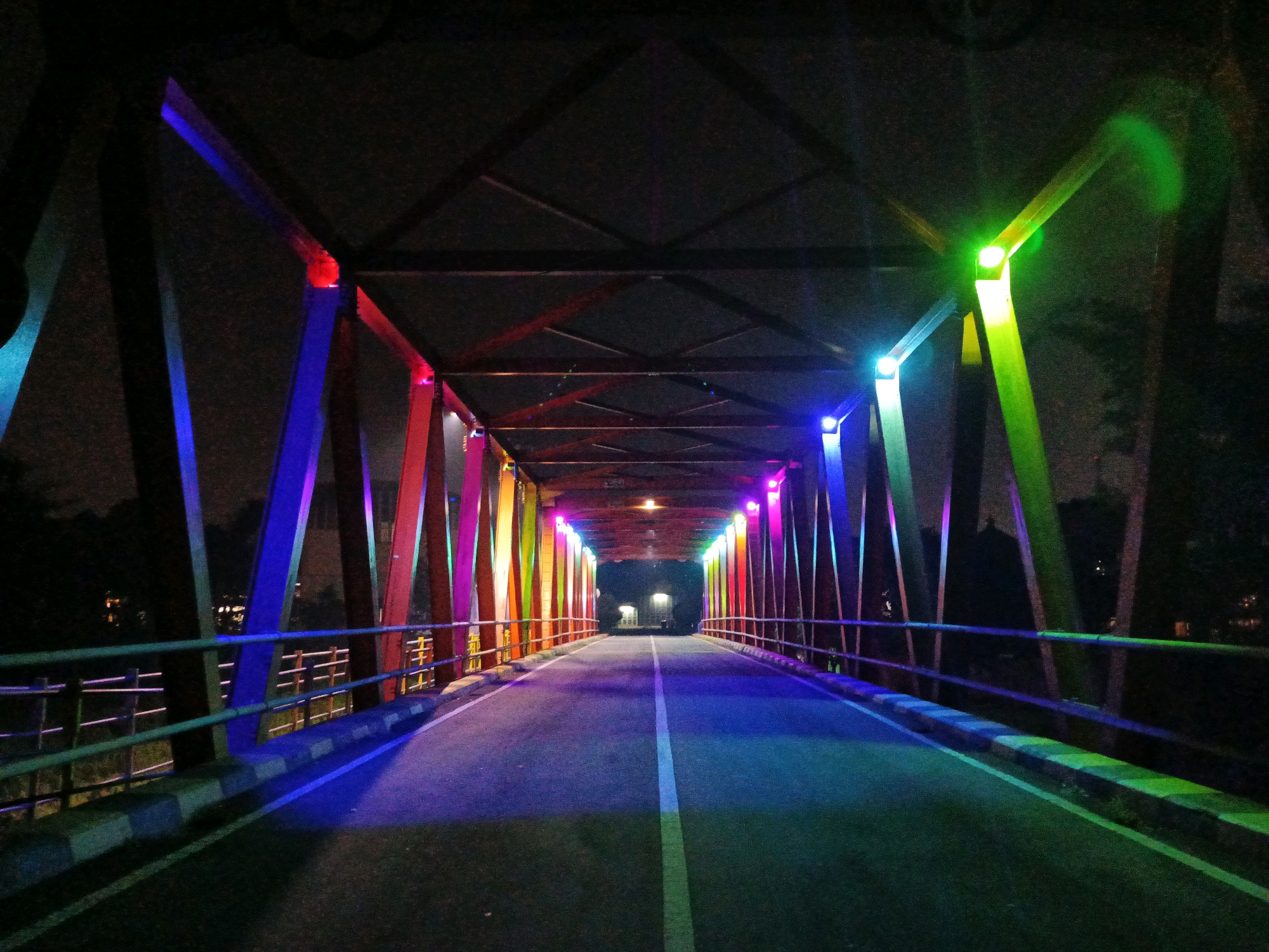 This is a nighttime photograph of a pedestrian bridge illuminated by colorful lights, likely in Bandung City, Indonesia. The bridge features a metal truss structure, with its vertical supports bathed in a spectrum of vibrant colors including blue, purple, pink, red, yellow, and green. The walkway of the bridge is paved with asphalt and has a white line dividing it down the center. Metal railings with horizontal bars line both sides of the walkway. The scene is dark, with the surrounding environment obscured by night. The lighting creates a dramatic and somewhat surreal atmosphere.