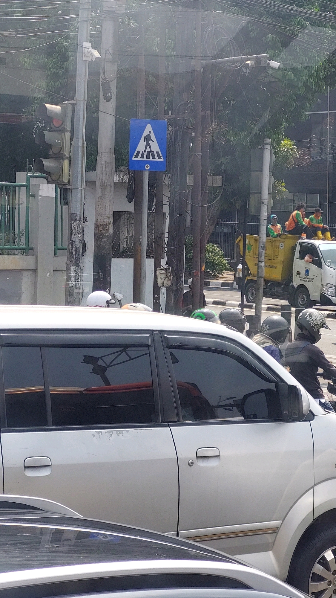 The image depicts a street scene in East Jakarta, Indonesia, on a bright day. In the foreground, the side of a silver vehicle is visible. Behind it, a road with several vehicles is present. To the left, a traffic light pole stands with a pedestrian crossing sign. The sign is a blue square with a white pedestrian silhouette walking across a white triangle, indicating a crosswalk. Several power lines and poles crisscross the upper portion of the image. Further in the background, a yellow truck with several people on it, possibly workers, is stopped at the side of the road. The environment appears to be urban, with buildings and trees lining the street. Several people are seen on motorcycles and walking, suggesting a busy street.