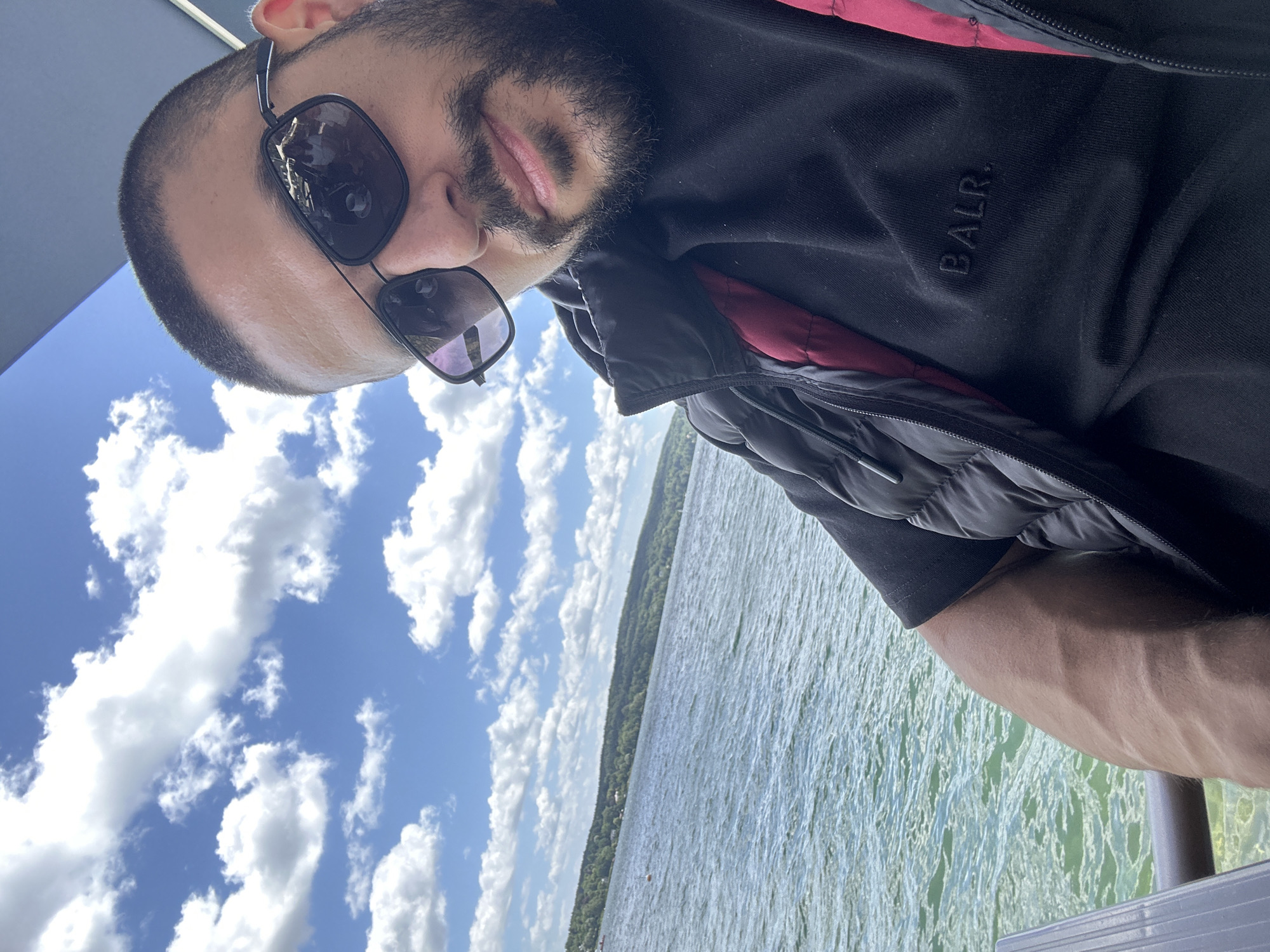 A man with short dark hair, a beard, and sunglasses is taking a selfie by a lake on a sunny day with a blue sky and white clouds. He is wearing a black puffer vest over a black t-shirt with the logo "BALR." visible on the front. The lake is visible in the background, with a tree-lined shore and some buildings in the distance. The image is taken from a slightly elevated angle, with the man's face in the foreground and the lake and sky stretching out behind him. The location is identified as the Starnberg Lake, Starnberg, Germany.