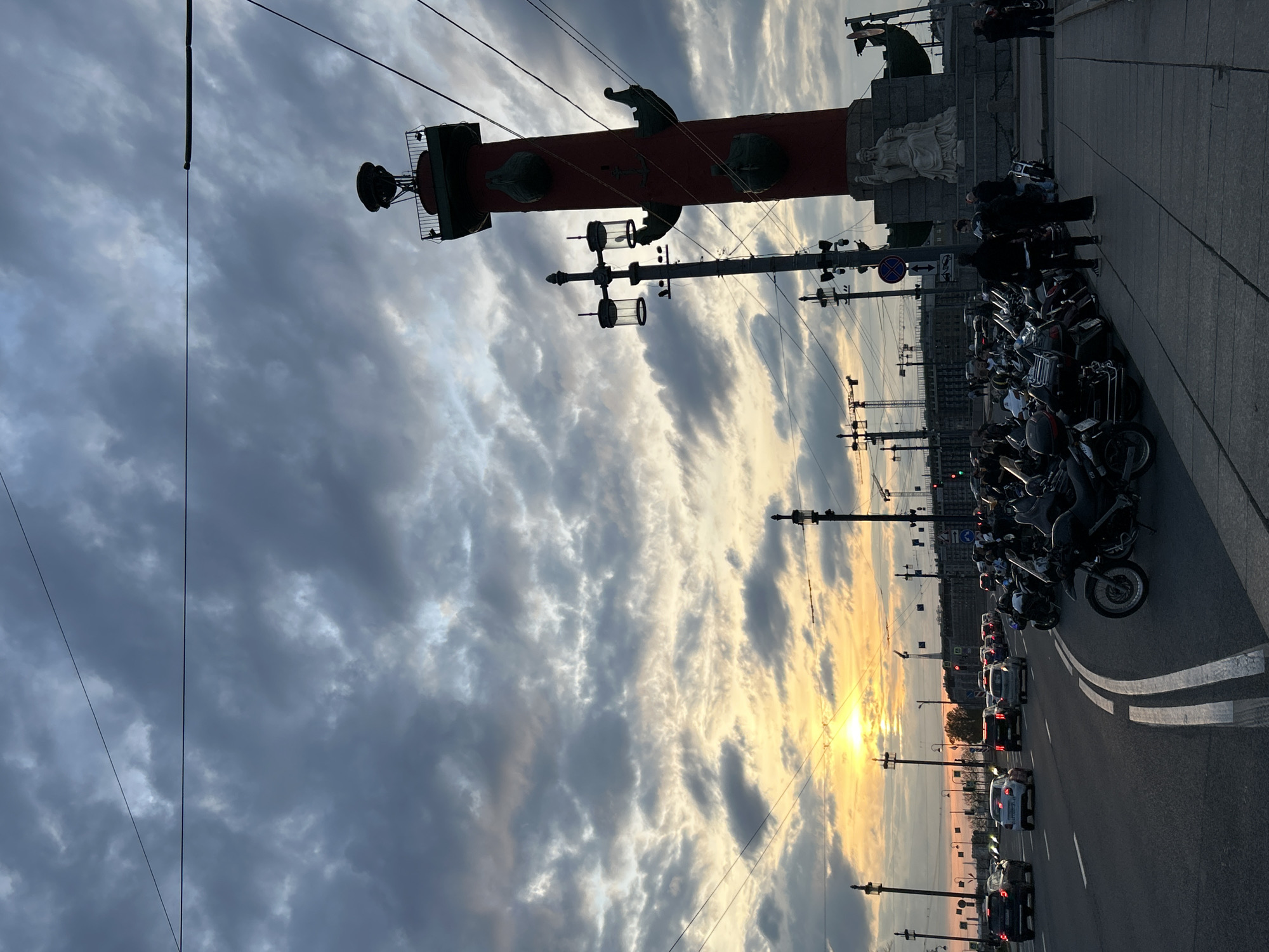 The image shows the Rostral Columns on the Spit of Vasilievsky Island in Saint Petersburg, Russia, at what appears to be sunrise or sunset. The sky is filled with clouds, and the sun is low on the horizon, creating a warm glow. The columns are red and topped with a decorative structure. Several motorcycles are parked on the sidewalk next to the monument. There is a busy road with cars driving. People can be seen standing near the monument. Overhead wires crisscross the sky.