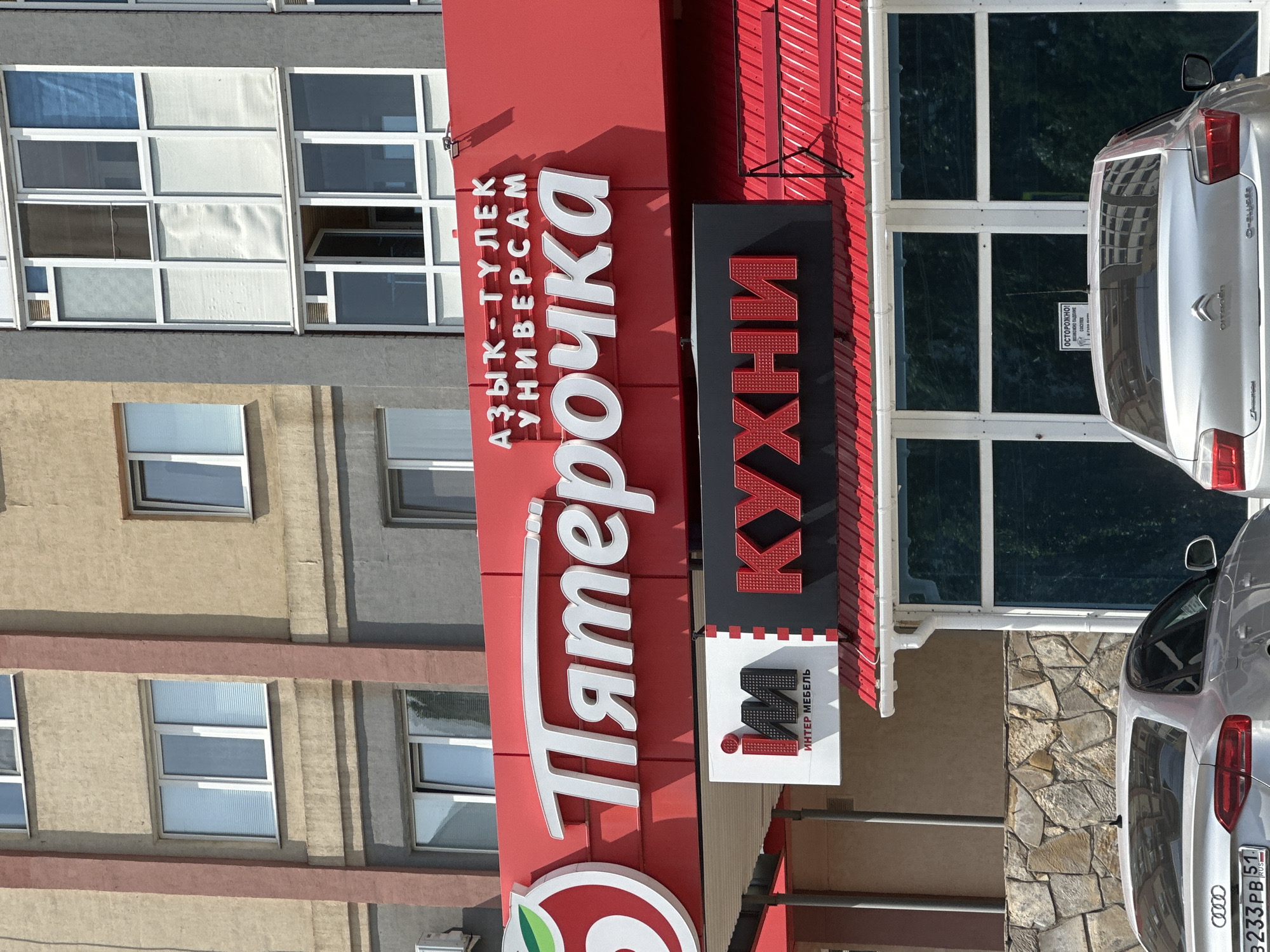 A daytime, eye-level, medium shot shows the exterior of a "Pyaterochka" grocery store in Sterlitamak, Russia. The store's prominent red signage features the white text "Пятёрочка" and above it, smaller white text reads "АЗЫК-ТУЛЕК УНИВЕРСАМ". To the left of the main signage is a circular green and white logo. Below the "Pyaterochka" sign, another sign in red and black displays the word "КУХНИ" (Kitchens) in large red letters, with a smaller black and white sign to its left that reads "INTER MEBEL" (Inter Furniture) in white text over a black 'im' logo. The building's facade above the signage is a combination of beige and gray, with multiple windows, some featuring white blinds. Below the store's awning, two silver cars are partially visible. The car in the foreground has a license plate reading "B233PB 51". The environment appears to be urban, with a clear sky overhead.