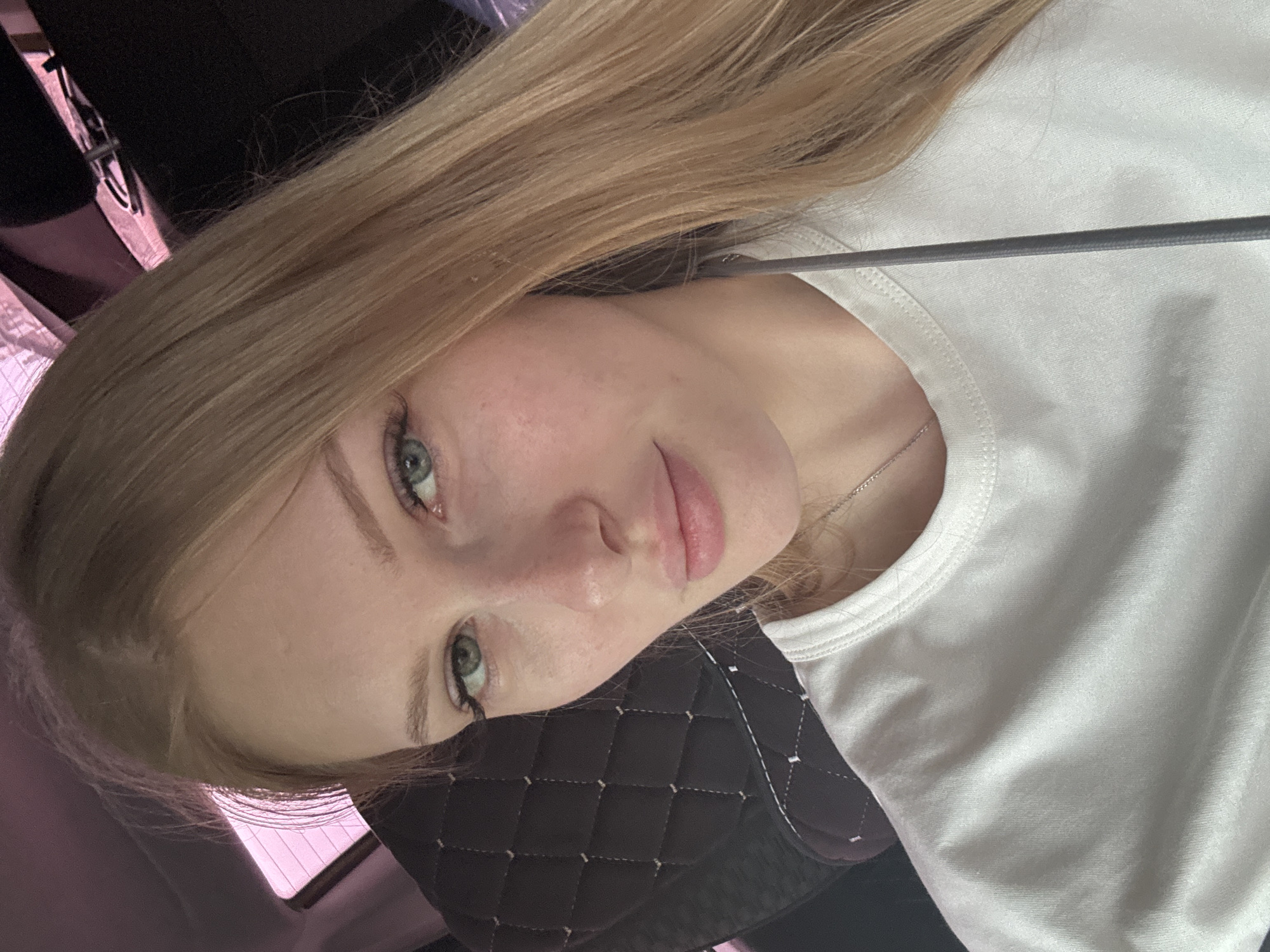 The image is a close-up, over-the-shoulder selfie of a young Caucasian woman with long, blonde hair and green eyes. She is wearing a white t-shirt and a thin silver necklace. Her makeup includes mascara and lipstick. The background appears to be the interior of a vehicle, with a quilted dark-colored seat visible on the left and a window with a pinkish tint on the upper left. A dark object, possibly a cable or strap, runs down the center of the frame. The lighting suggests it is daytime. The location is specified as the city of Perm, Russia.