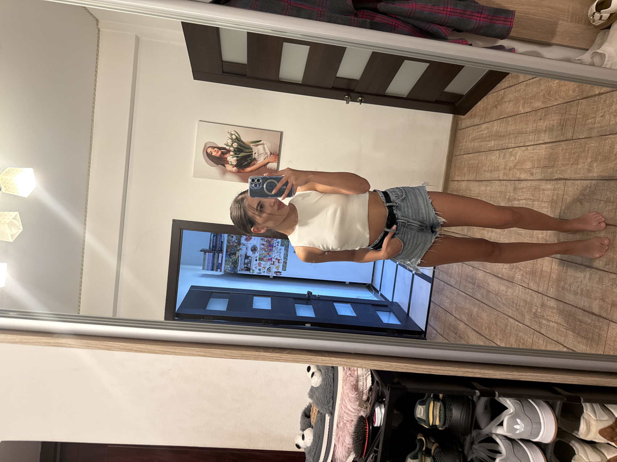 In an indoor setting, a young woman stands facing a large mirror, taking a selfie with her phone. She is wearing a white cropped tank top and denim shorts. Her reflection is visible in the mirror, showing her full body and the surrounding environment. Behind her, a dark-colored door is partially open, revealing a glimpse of what appears to be a hallway with tiled flooring. To her right, a plaid shirt hangs on a hook. To her left, a shoe rack is filled with various footwear, including sneakers and slippers. In the reflection, a framed picture of a woman wearing a hat and holding flowers is visible on the wall. The floor is made of wooden planks. The scene suggests a private residence or dressing area.