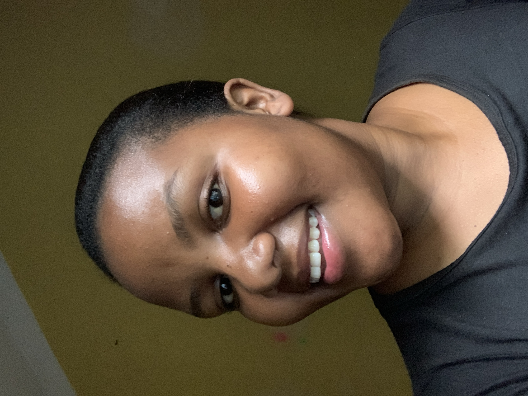 A close-up portrait of a woman with dark skin and short, dark hair, angled slightly to the side. She is smiling, revealing white teeth and pink lips. Her eyes are dark and appear moist. Her skin is smooth and appears to have a slight sheen. She is wearing a black top. The background is a solid, muted yellow color. There is no specific evidence to suggest that the photograph was taken in Dar es-Salaam, Tanzania.