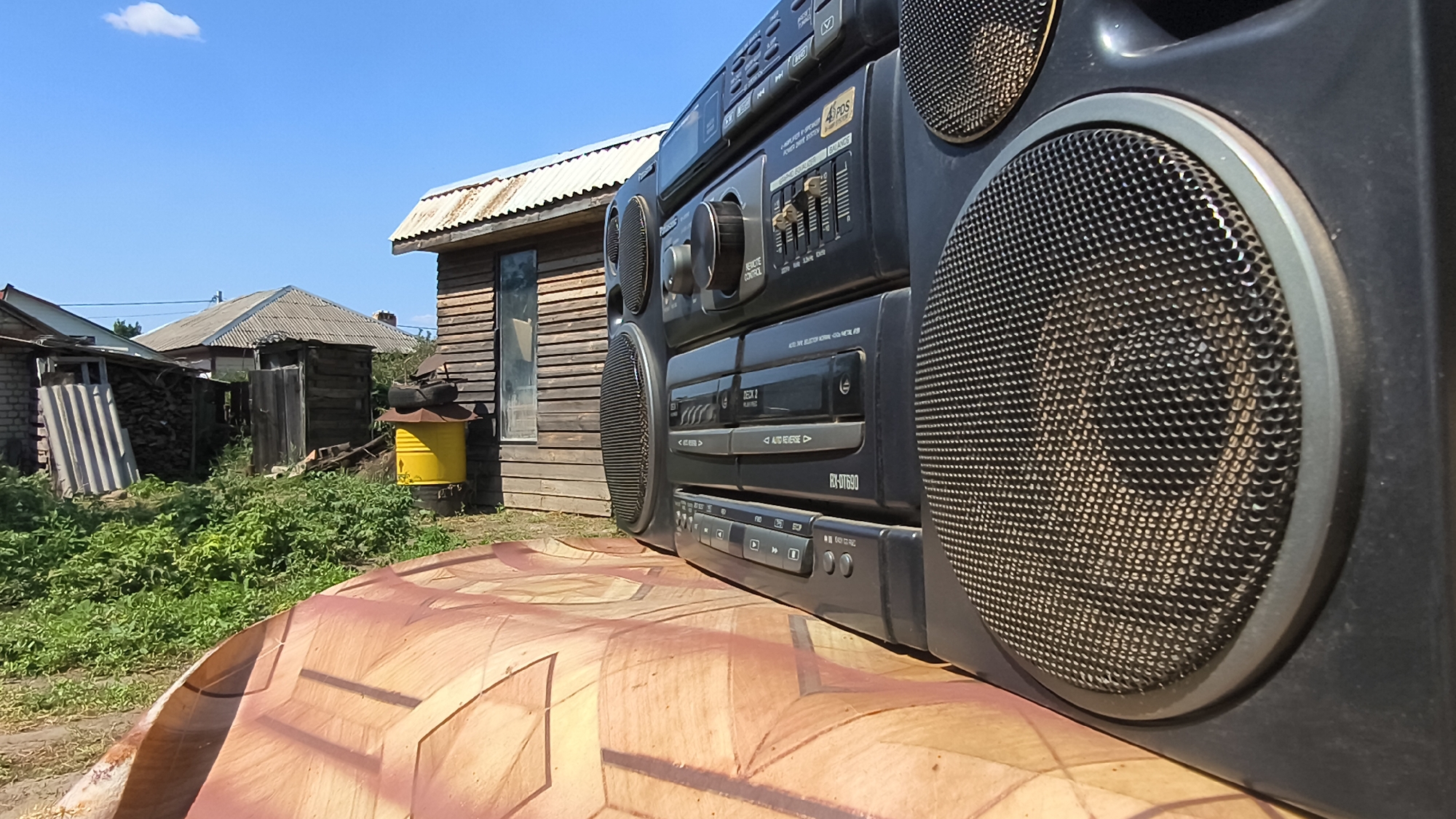 An outdoor, medium shot captures a black boombox in the foreground, positioned on a surface with a brown, textured pattern. The boombox, which appears to be a Panasonic RX-DT690 model, is angled with its front controls and speakers visible. Behind the boombox, the scene depicts a rustic, rural setting. Several wooden structures, possibly small houses or sheds with corrugated metal roofs, are visible. A bright yellow barrel stands near one of the wooden buildings, surrounded by dense green vegetation. The sky above is clear and blue with a single white cloud. The overall environment suggests a sunny day in a village or a suburban area with a somewhat unkempt appearance, possibly in Mikhaylovka, Russia. There are no people or animals in the frame.