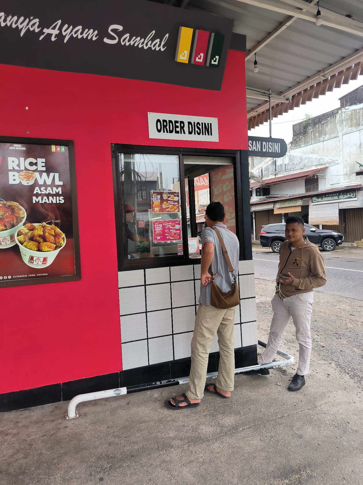 An outdoor food kiosk with a red and black exterior is depicted, featuring a large advertisement for "RICE BOWL ASAM MANIS" on the left. Above the window, a sign reads "ORDER DISINI" (Order Here). Two men are present: one in a grey t-shirt and khaki pants, wearing a brown crossbody bag, stands at the order window. The second man, in a light brown hooded jacket and light-colored pants, stands to the right, holding a smartphone. In the background, a dark SUV is parked on a street lined with buildings. The scene appears to be in Meulaboh, Indonesia, as suggested by the style of architecture and signage.