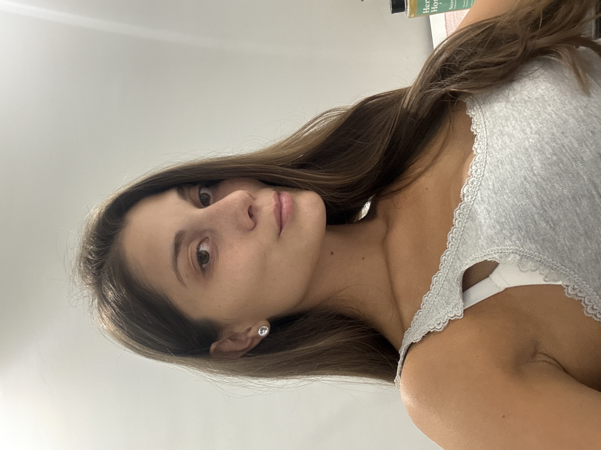 A young woman with brown hair is facing the camera. She is wearing a grey tank top with lace trim. The background is a plain white wall. The image was not taken in Burgas, Bulgaria.