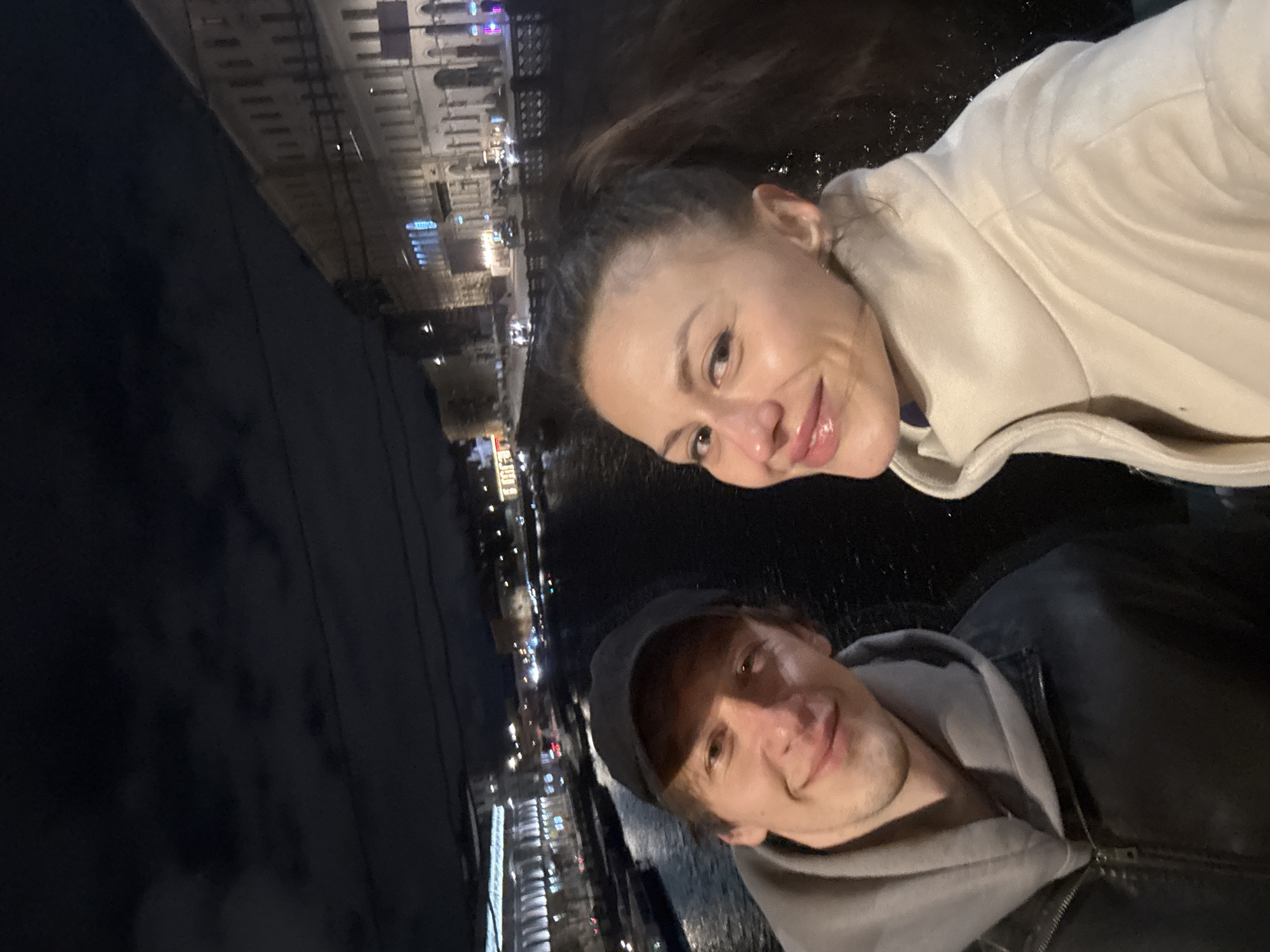 Two individuals, a woman and a man, are positioned closely together, suggesting a selfie or a photo taken at close range. The woman is slightly above the man, with her face appearing more prominently. She has fair skin, dark hair pulled back in a ponytail, and is wearing a light-colored coat. The man has lighter hair and is wearing a cap and a jacket with a hood.
Behind them is a body of water, presumably a river or canal, with illuminated buildings visible along the waterfront. The buildings suggest an urban setting. The location is Saint Petersburg, Russia.