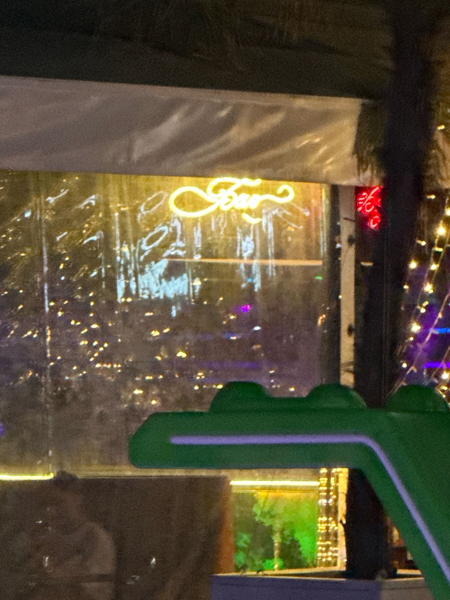 The image depicts an outdoor scene at night, likely a bar or restaurant with outdoor seating in Lermontovo, Russia. In the foreground, a large, green, stylized crocodile-shaped object is visible, with purple accents along its edge. Behind this object, a transparent plastic sheeting hangs, suggesting it's a temporary enclosure for an outdoor area. Through the sheeting, a neon sign in a cursive font, appearing to spell "Bar" in yellow, is prominently displayed. Scattered lights, some appearing as bokeh effects, are visible in the background, indicating a city environment or decorative lighting. To the right of the "Bar" sign, a smaller red neon sign with what appears to be Cyrillic characters is present. Further to the right, a string of fairy lights, some purple and some white, are illuminated, suggesting a festive atmosphere. In the lower left quadrant, a blurred human figure can be seen sitting, possibly a patron of the establishment. The overall scene suggests a lively evening setting, possibly a waterfront or entertainment district, with the presence of the crocodile object hinting at a playful or themed establishment.