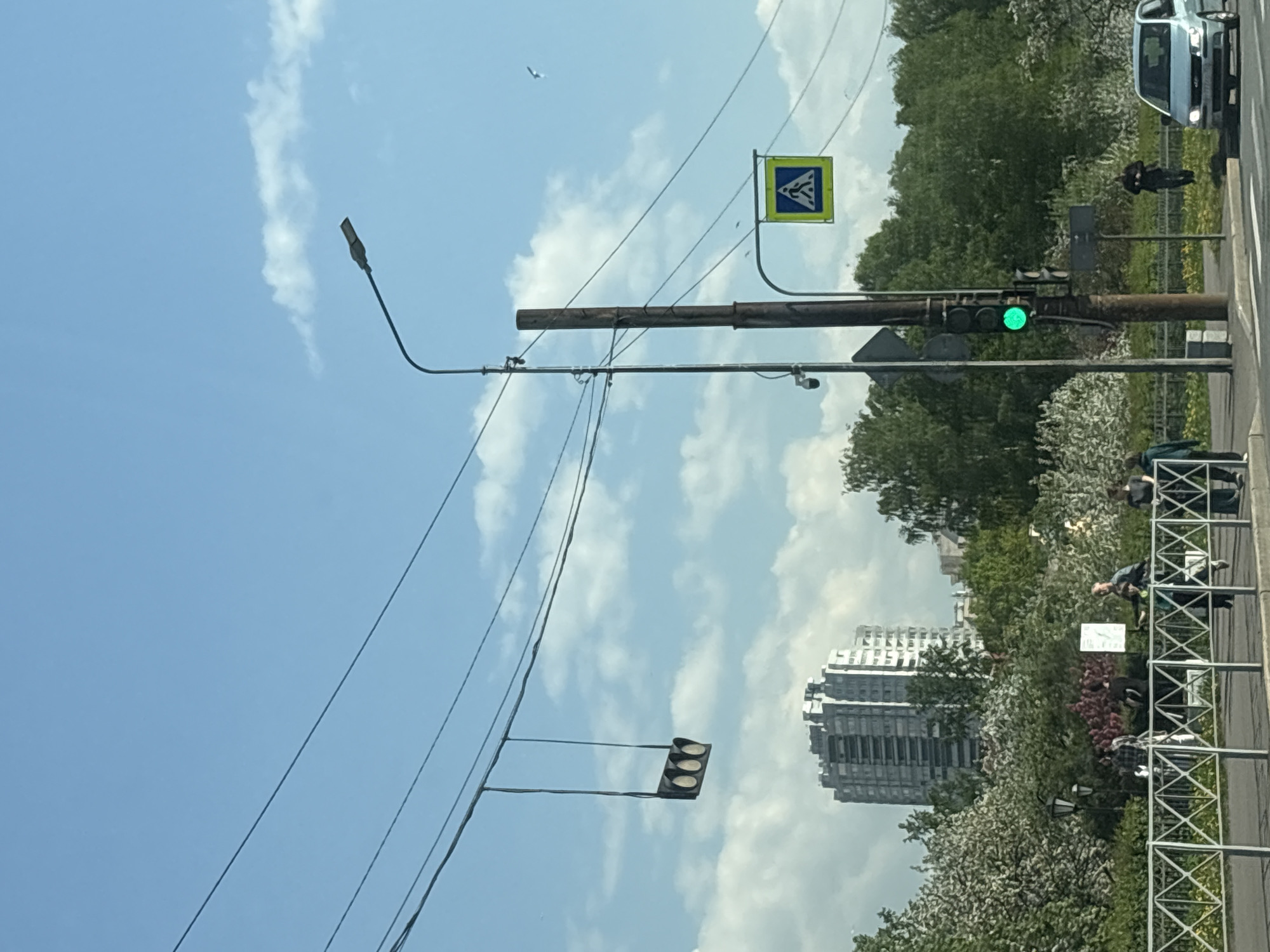 The image depicts a street scene in Saint Petersburg, Russia. The sky is partly cloudy. A traffic light is green. There is a street light and pedestrian crossing sign. People are on the sidewalk near a metal barrier. A white van is at the far right of the frame. A tall building is visible in the distance behind trees.