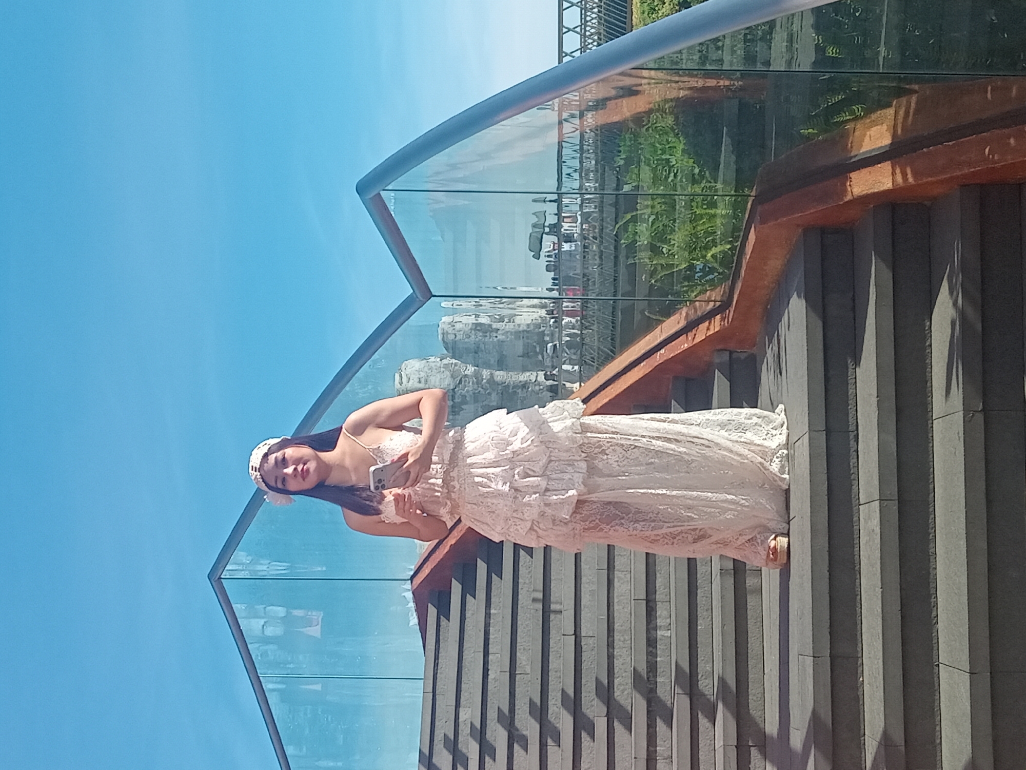 A young woman is captured in a vertical, full-body shot standing on a modern outdoor staircase with glass railings. She is wearing a white, floor-length lace dress with ruffles and a white headpiece. Her hair is dark and cascades over her shoulders. She is holding a smartphone in her right hand. The staircase is constructed with dark, rectangular stone steps and has a reddish-brown wooden trim along the edges. The glass railings offer a reflective view of lush greenery and some architectural elements.
The scene appears to be set in a sunny outdoor location, indicated by the bright blue sky and the sharp shadows cast on the stairs, suggesting it is daytime, likely late morning or early afternoon. The presence of plants and the modern architectural design of the staircase hint at a contemporary public space, possibly a park, a viewpoint, or a stylish urban development. The woman's pose and attire, particularly the delicate white dress, suggest a formal or celebratory occasion, or perhaps a staged photoshoot. Her facial expression is relaxed, with a slight smile, and she seems to be looking towards the camera. No visible text is present in the image. The overall impression is one of serene elegance against a backdrop of contemporary design and natural elements.