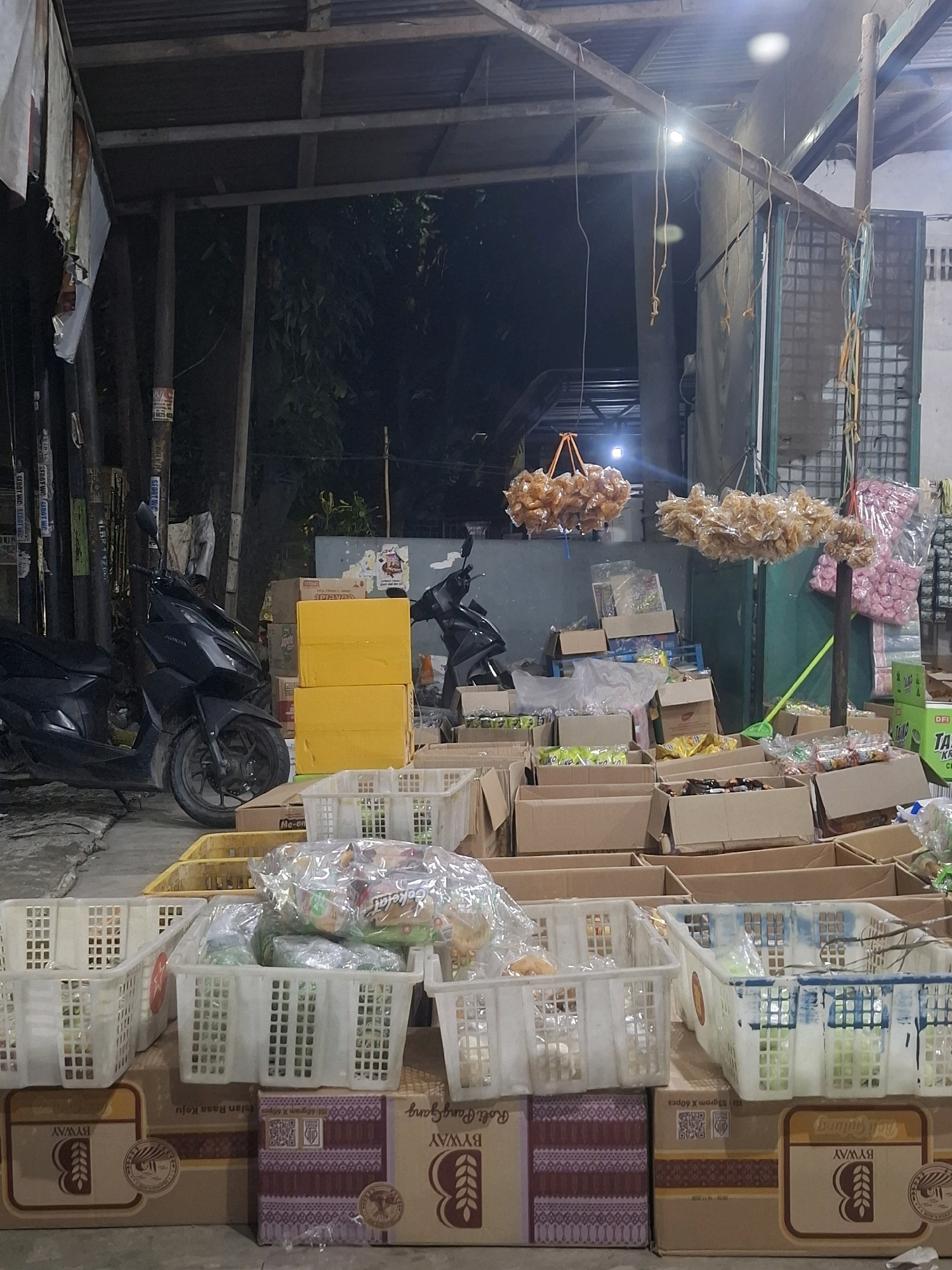 The image depicts an outdoor market stall in the city of Medan, Indonesia, likely at dusk or night, given the artificial lighting. The stall is cluttered with various goods, predominantly stacked cardboard boxes and plastic crates. The boxes are arranged in multiple tiers, some open and revealing packaged items inside, while others remain sealed. The plastic crates, mostly white and rectangular with grid patterns, are also filled with packaged goods.
Several hanging items are visible: two clusters of what appear to be snack foods or dried goods are suspended from a wooden beam overhead. One cluster is a light brown, while the other is a pale yellow. To the right, a large pink cluster of similar items is partially visible.
A black scooter is parked to the left of the frame, partially obscuring some of the boxes. In the background, more structures are visible, including what seems to be a corrugated metal roof and wooden supports. The overall impression is one of a busy, perhaps slightly disorganized, retail space. The ground appears to be a paved surface. No people are clearly visible in the frame.