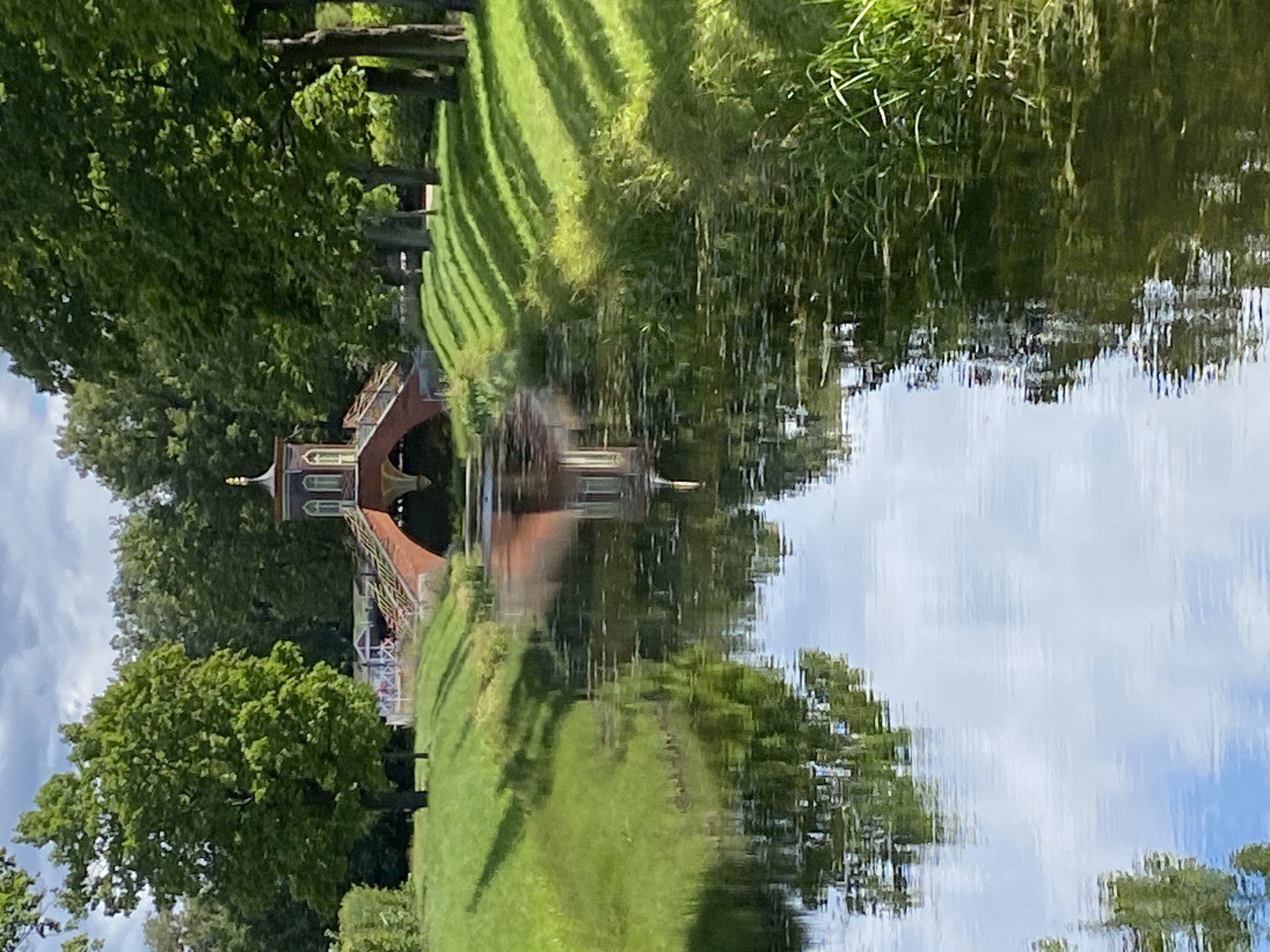 This is a medium-distance, eye-level, outdoor, vertical shot of a scenic canal in the Catherine Park, located in the city of Pushkin, Russia. The canal flows from the foreground towards a decorative brick bridge in the background. Lush green grass covers the banks of the canal, with trees lining the right bank, casting dappled shadows on the grass. The water is calm and reflects the sky, trees, and the bridge. The bridge is ornate, with a central pavilion topped by a golden finial. The scene is peaceful and idyllic, typical of a park setting. There are no people or animals visible in the frame.