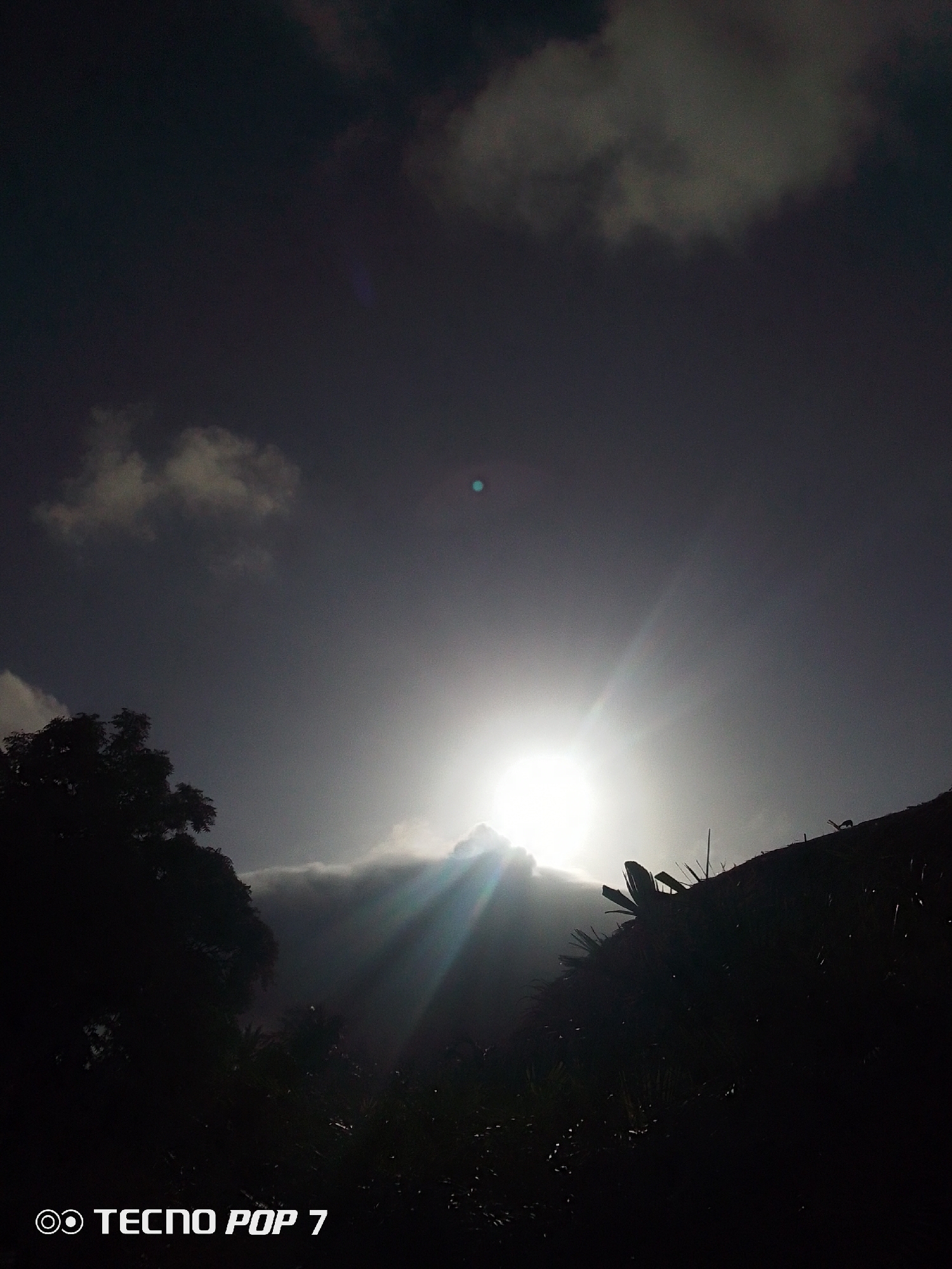 The image shows a sunburst through clouds in what appears to be a landscape with trees and other vegetation in silhouette. The sky is dark, and the sun is low on the horizon. The image was taken with a "TECNO POP 7" camera. The location is identified as Pedro González, Venezuela.