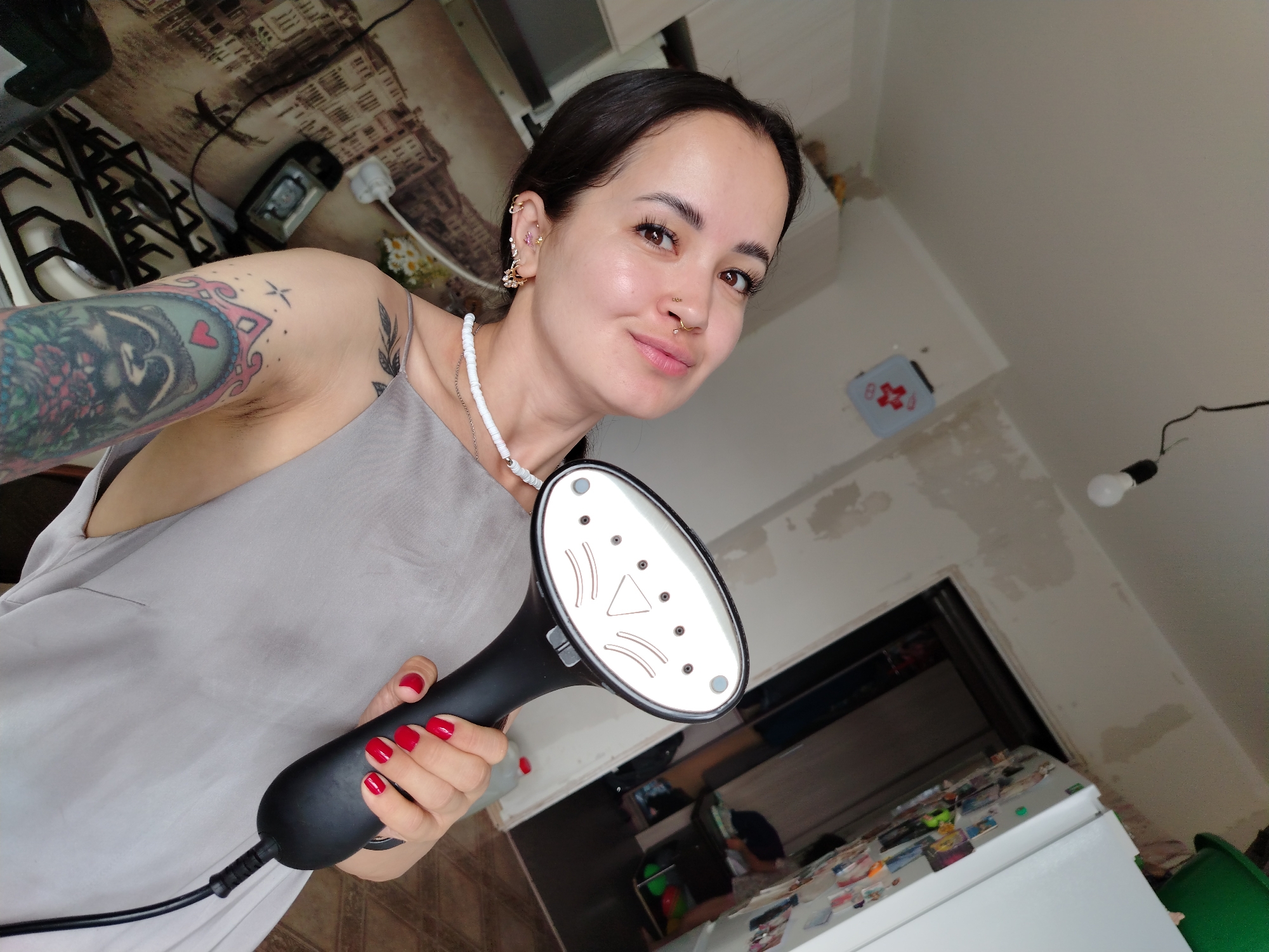 A woman with dark hair, brown eyes, and multiple piercings on her ears and nose is taking a selfie in what appears to be a kitchen in Noviy, Russia. She is wearing a gray sleeveless top and has a large tattoo on her left arm. She is holding a black handheld garment steamer with a white soleplate, and her fingernails are painted red. The background shows a gas stove, a refrigerator, and partially painted walls, suggesting the area might be undergoing renovation or is in a state of disarray.