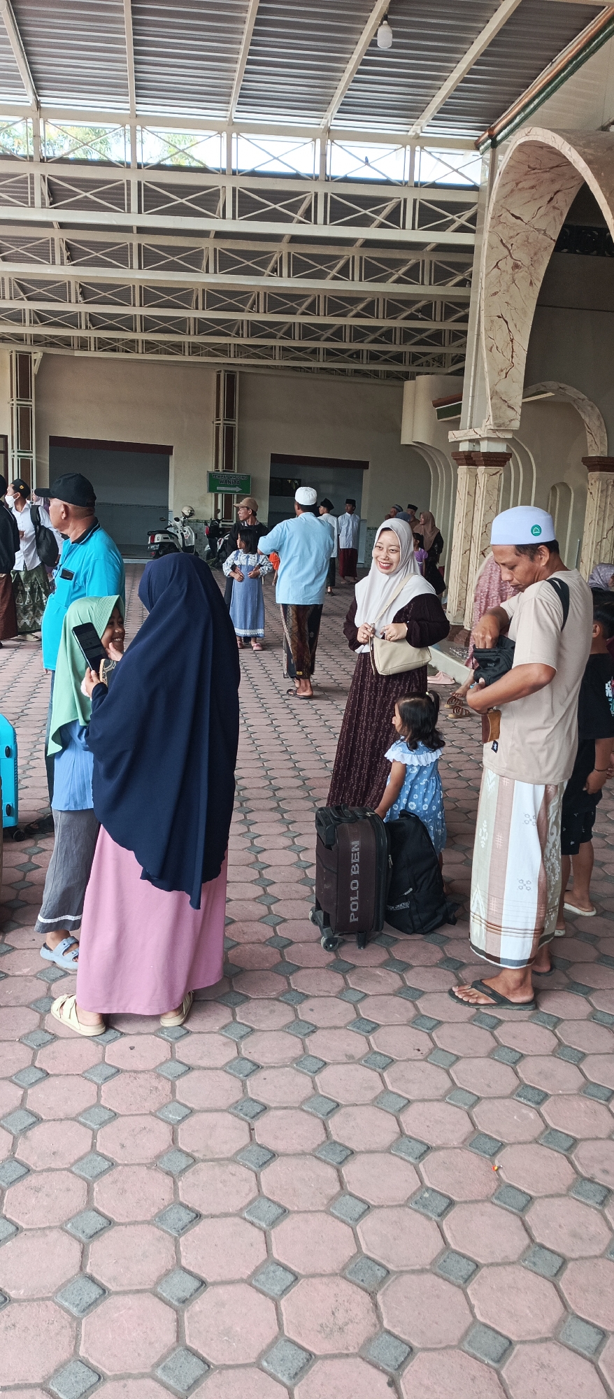 In Kapongan, Indonesia, an outdoor scene unfolds under a large, slatted roof structure, supported by a white, industrial-style frame. The ground is paved with red and grey octagonal and square tiles. A group of people, predominantly dressed in traditional Muslim attire, are gathered, suggesting an event or travel.
In the foreground left, a person in a teal shirt and a black cap stands next to a person in a navy blue headscarf and a long pink skirt. The person in the navy headscarf holds a smartphone, appearing to take a photo. To their right, a woman in a patterned brown and white dress and a light beige headscarf smiles towards the camera, holding a beige purse.
Further back, a man in a light blue long-sleeved shirt and patterned trousers is interacting with a young girl in a blue dress with white ruffles. Beside them is a black rolling suitcase with "POLO BEN" written on it, and a black backpack.
In the background, several other individuals are visible, some in white robes and skullcaps, and a motorcycle is parked near an entrance labeled "TEMPAT WUDHU WANITA" (Women's Ablution Area). The overall atmosphere suggests a communal gathering, possibly before or after a religious service or a journey. The architecture of the roof and the patterned tiling of the ground indicate a public space, likely a mosque complex or a transportation hub.