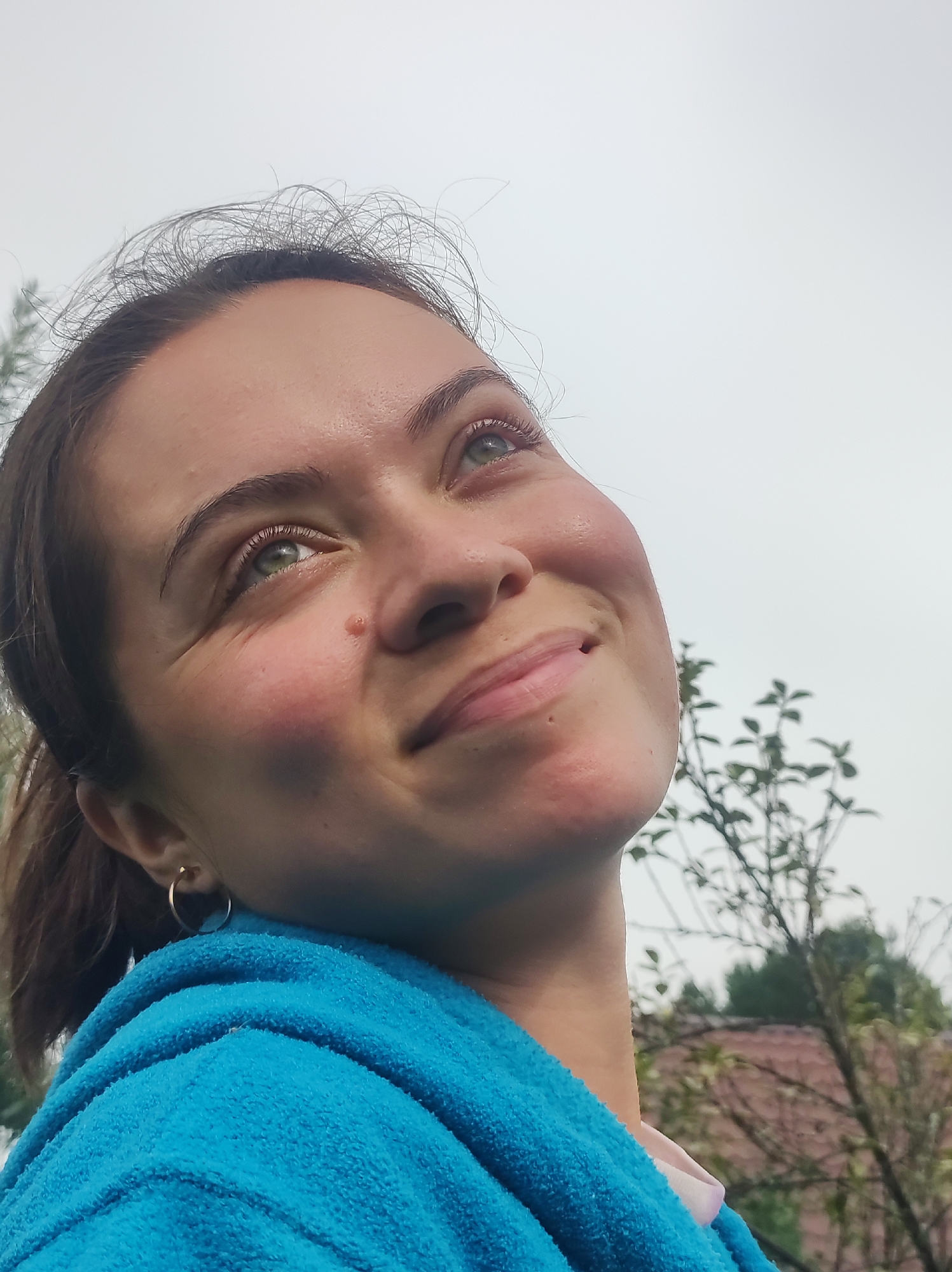 A close-up, low-angle shot captures a young woman with light brown hair and green eyes looking upwards and to her right, a slight smile on her face. Her skin appears fair with rosy cheeks. She is wearing a bright blue towel draped around her shoulders, and a small gold hoop earring is visible on her left ear. The background is an overcast sky with some blurred green foliage and tree branches, suggesting an outdoor setting, potentially in the city of Fokinsky, Russia, given the provided context. The image is well-lit, with the light source appearing to be from above.